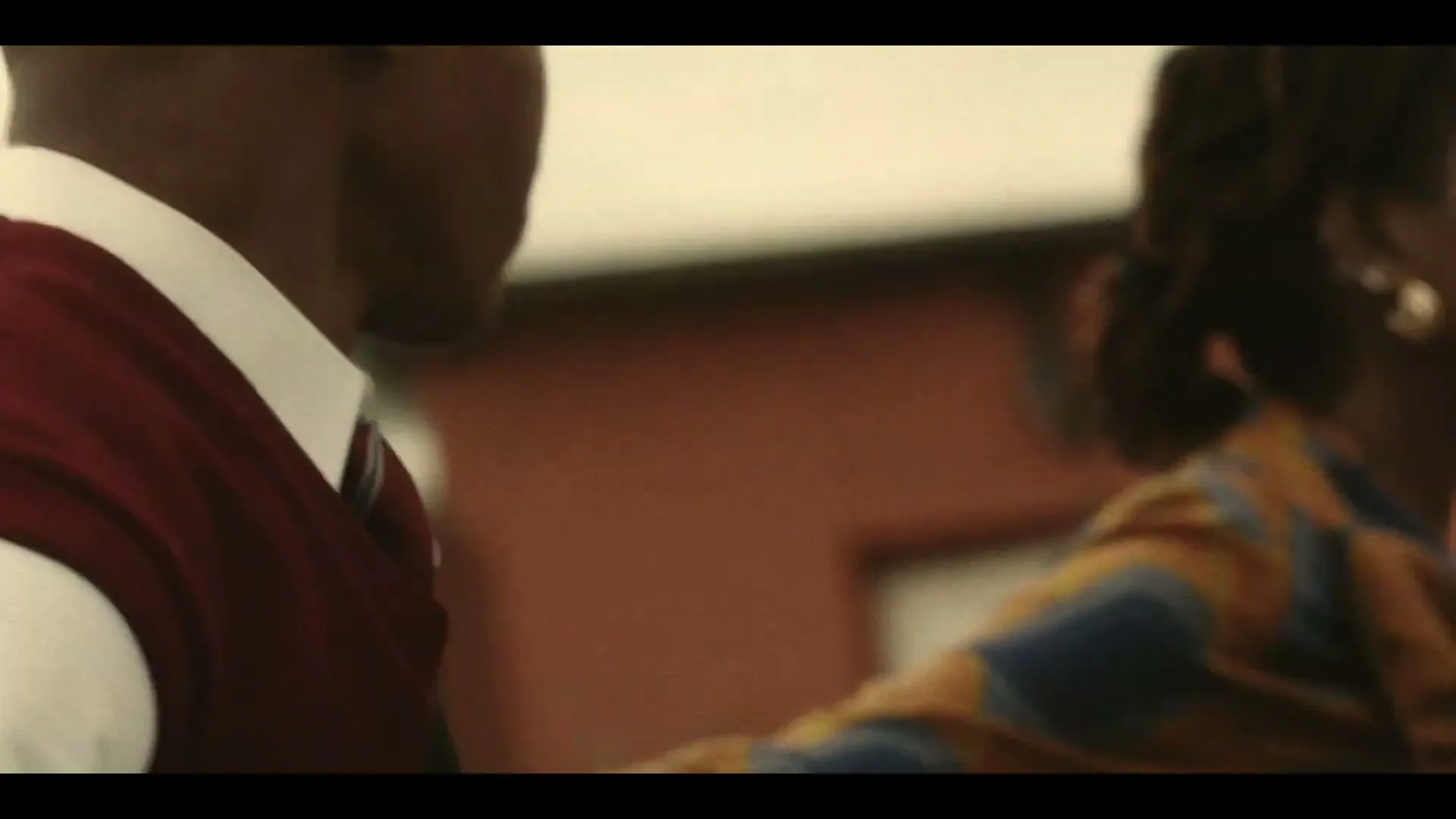A young Black male student in a school uniform looks off-screen, with another person partially visible beside him, in an outdoor setting.