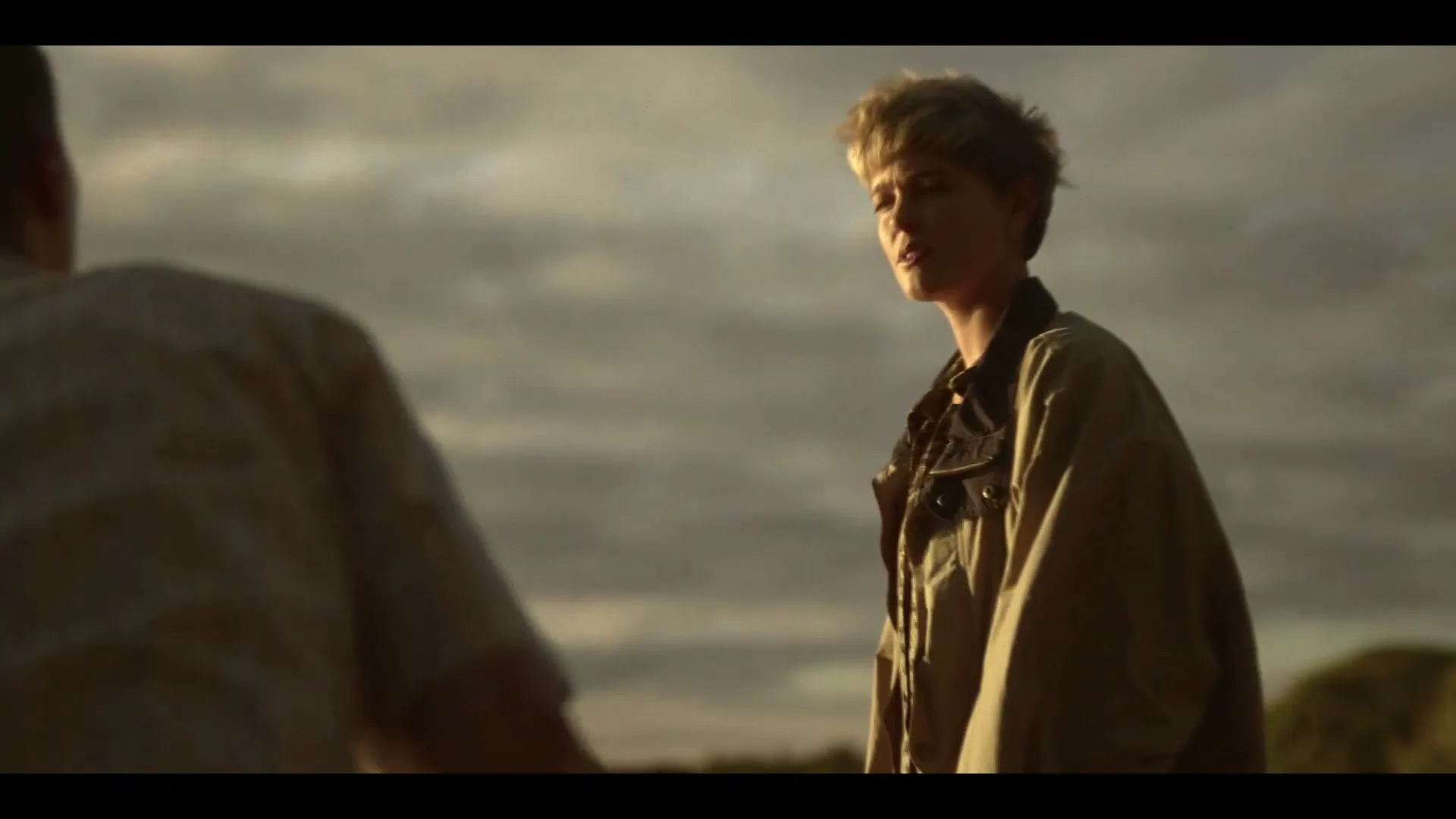 A young woman with short blonde hair and a tan jacket looks intently at a man with his back to the camera, under a cloudy sky.