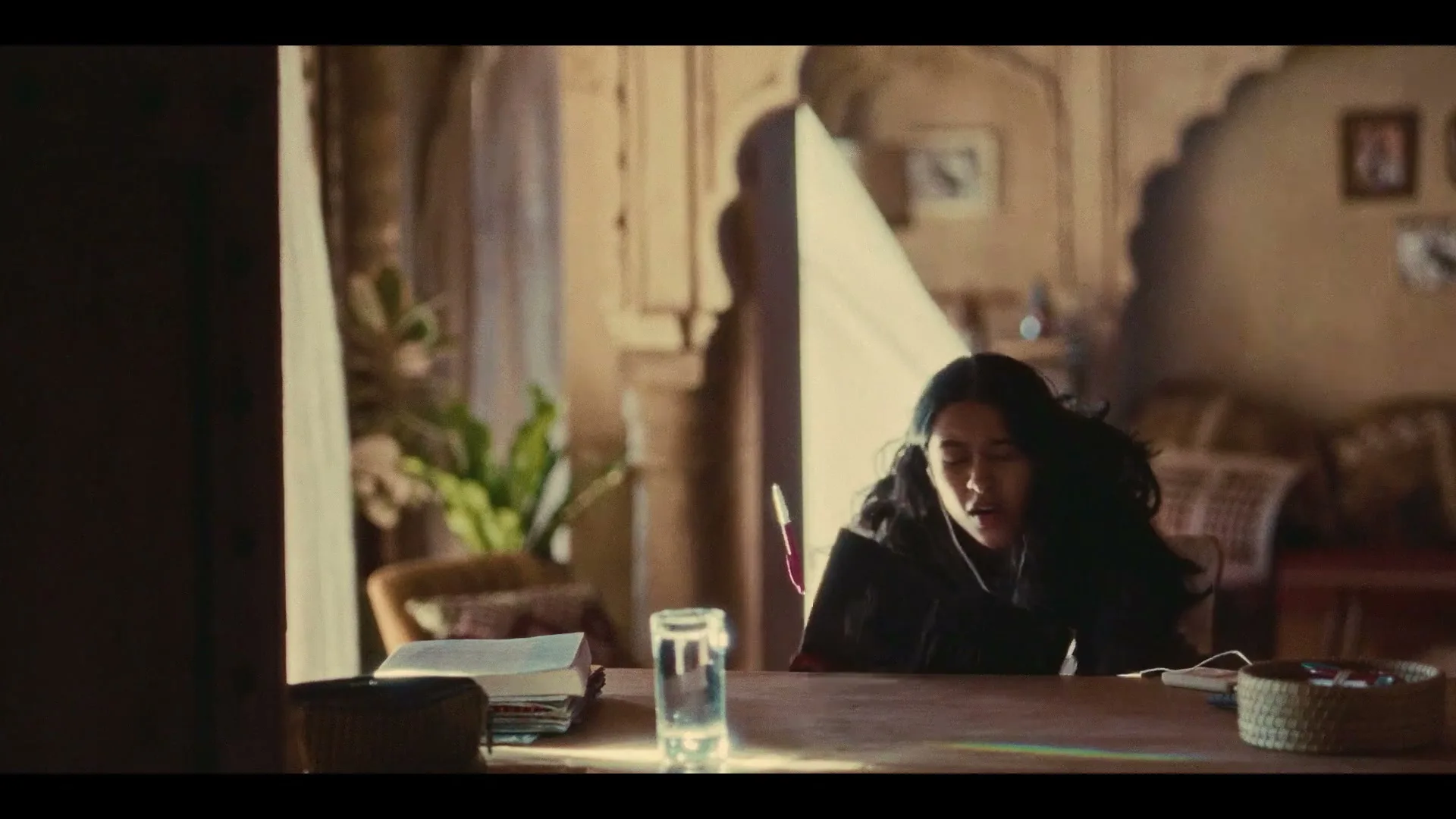 A young woman with long dark hair, wearing a dark top, sits at a wooden table, writing in a notebook in a sunlit room.
