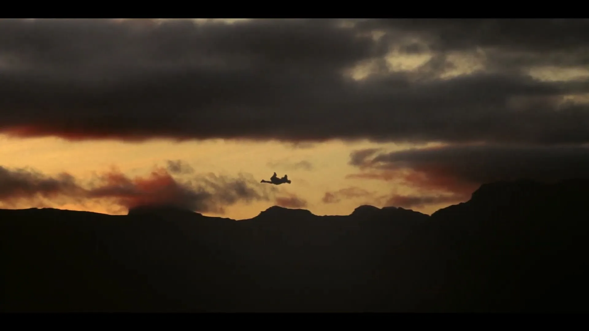 A silhouette of a person falls through the air against a dramatic sunset sky with dark clouds and mountains in the background.