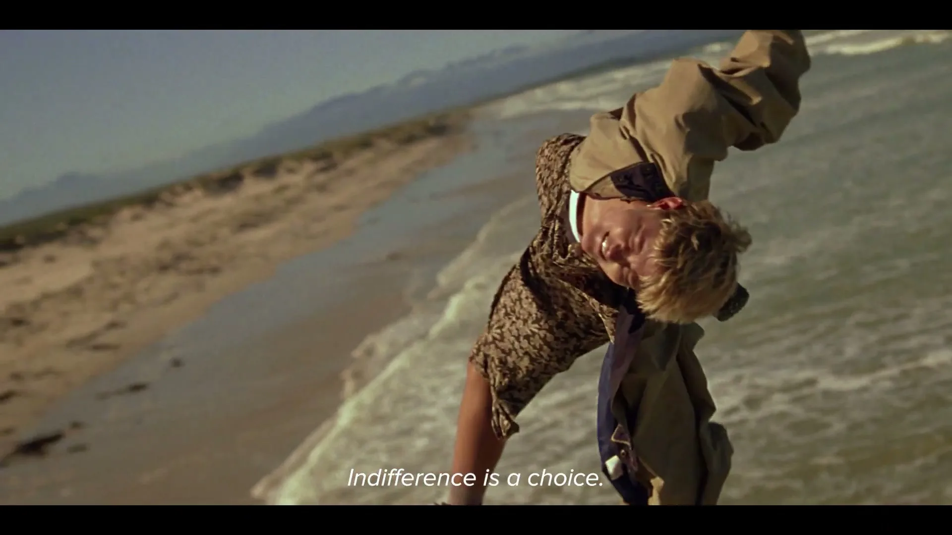 A young man with blonde hair, wearing a patterned shirt and jacket, hangs upside down over a beach with waves crashing, looking distressed.