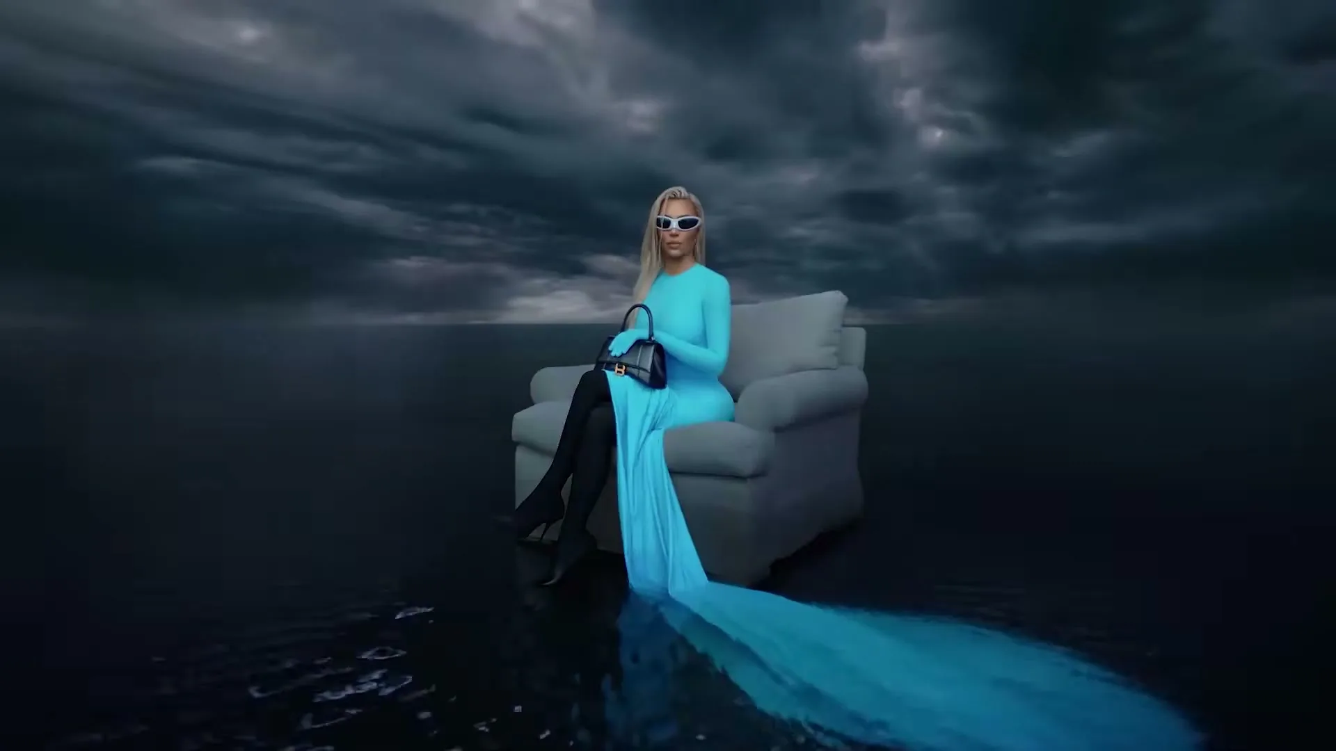 A blonde woman in a long blue dress and sunglasses sits on a couch in a dark, watery landscape under a stormy sky, holding a black handbag.