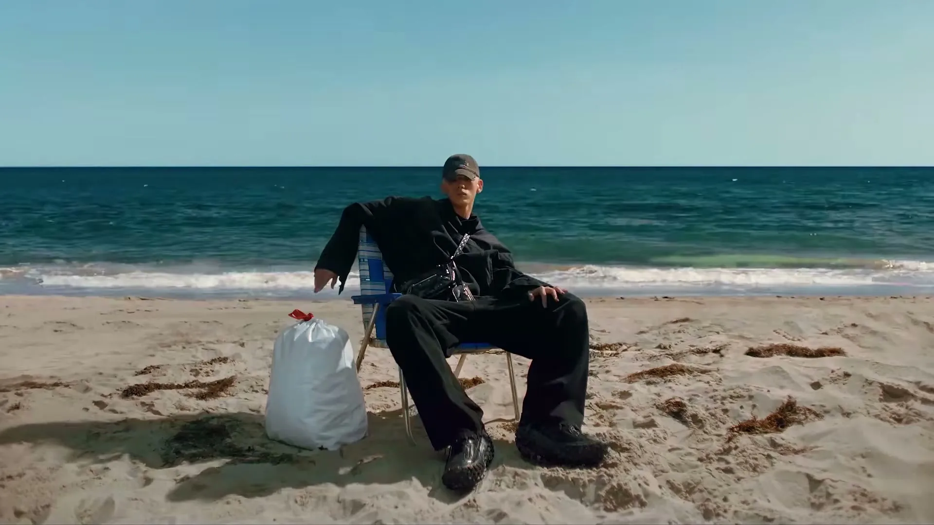 A young man in all black sits on a beach chair next to a white trash bag, looking out at the ocean on a sunny day.