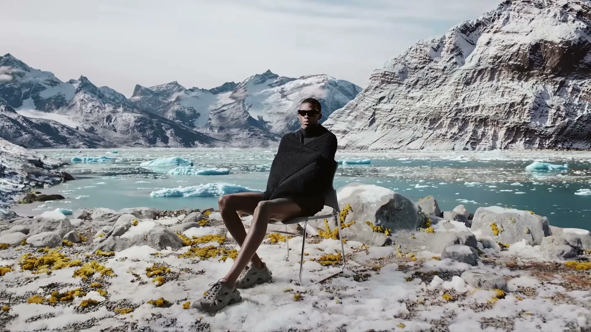 A model in a black poncho and shorts sits on a chair in a snowy, rocky landscape with a glacial lake and mountains in the background.