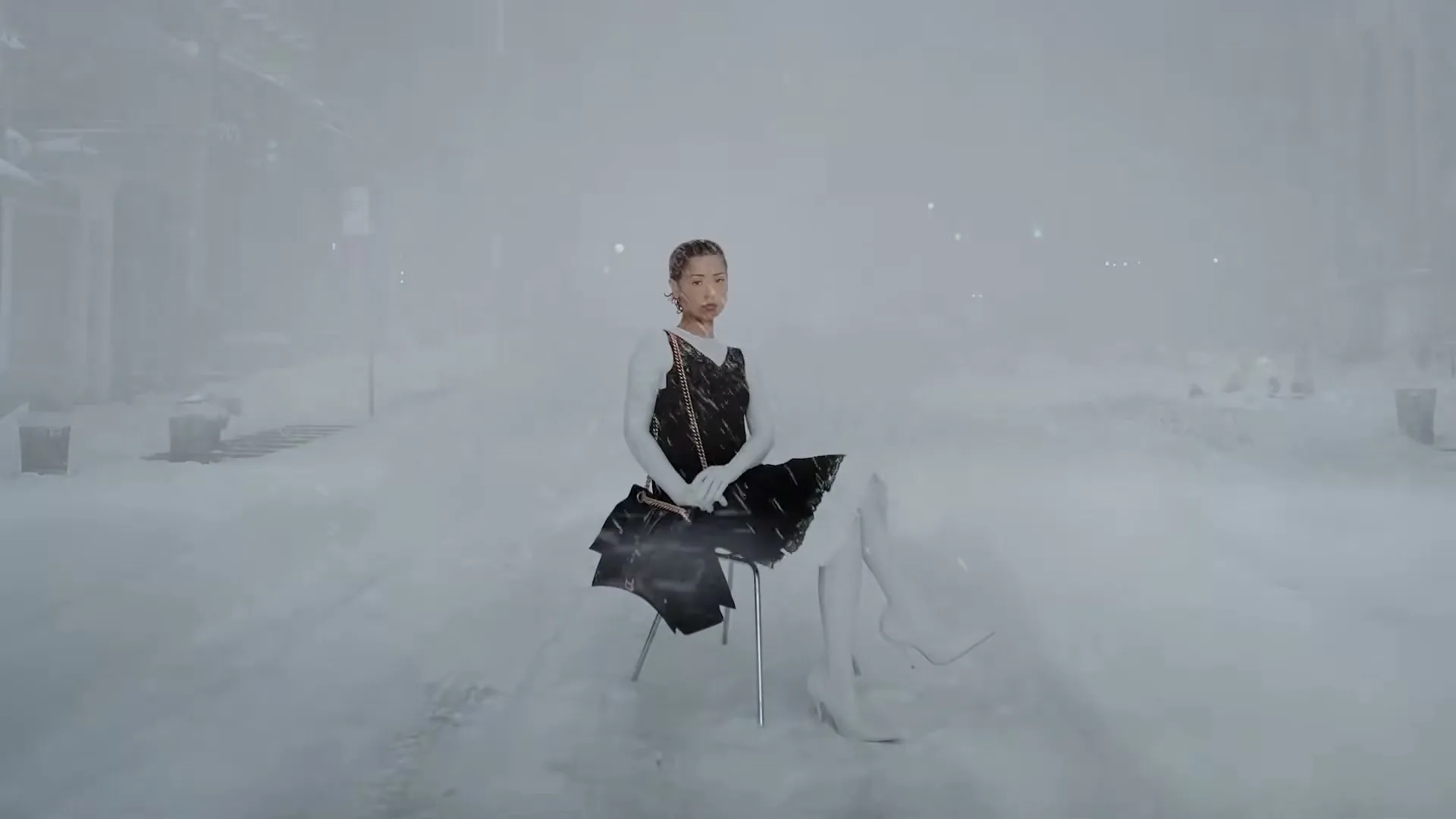 A female model in a black dress and white boots sits on a chair in the middle of a snow-covered street during a blizzard.