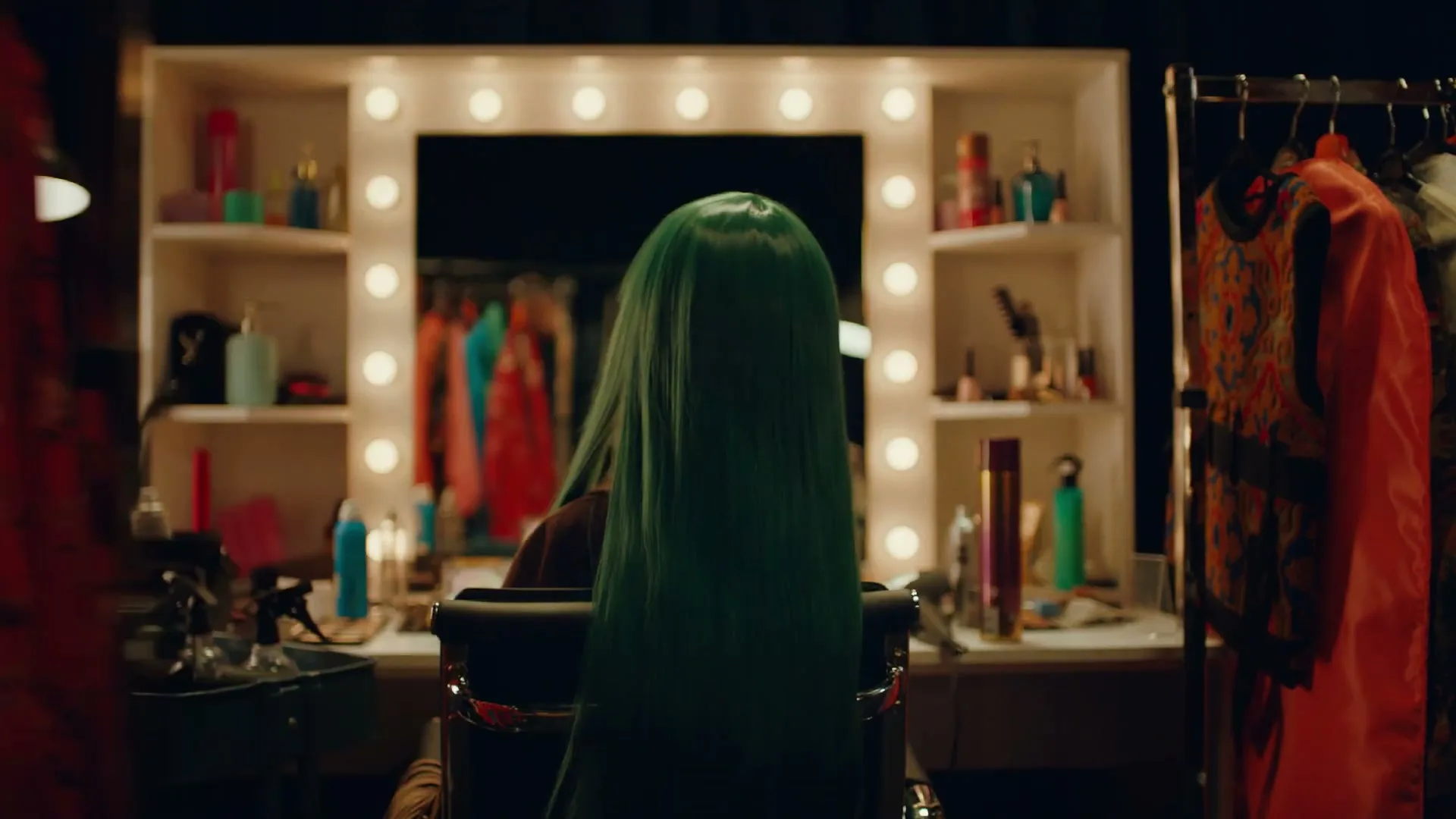 A person with long green hair sits at a brightly lit vanity mirror, surrounded by clothing racks and beauty products in a dressing room.