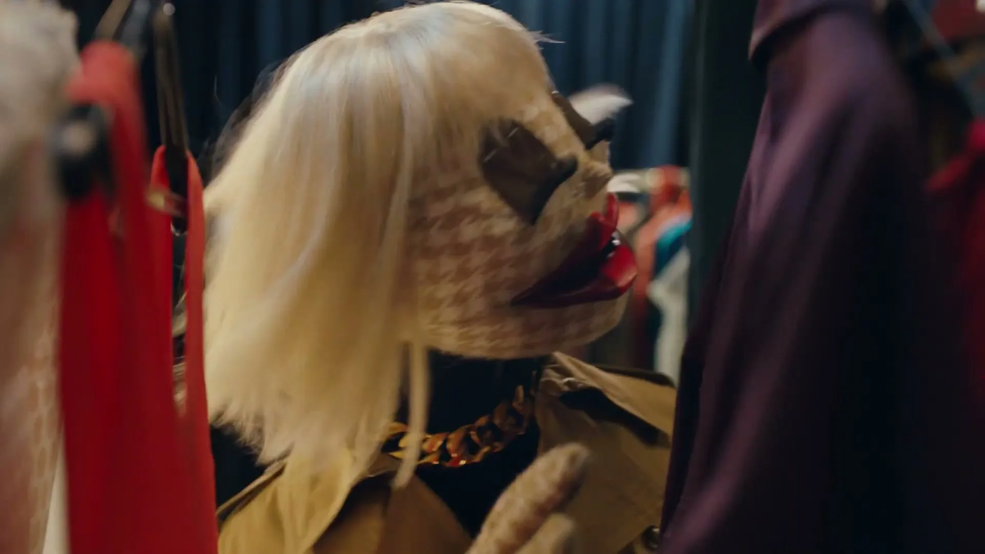 A puppet with a houndstooth face, blonde wig, and trench coat turns its head in a clothing rack.