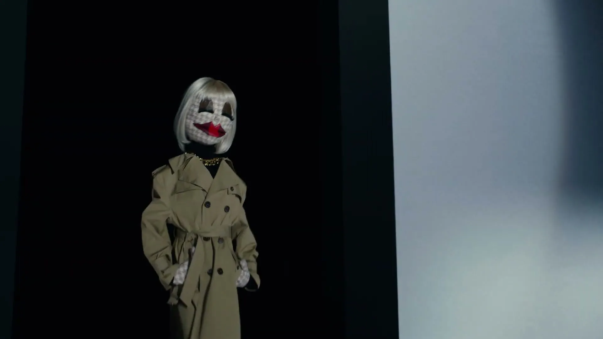 A puppet-like figure with a large head, blonde hair, and red lips, wearing a beige trench coat, stands against a dark background.