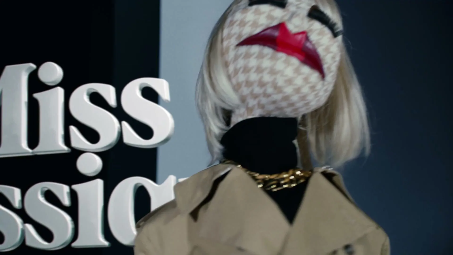 A puppet with a houndstooth face, blonde hair, and red lips wears a trench coat and gold chain, turning its head to the right in a studio.