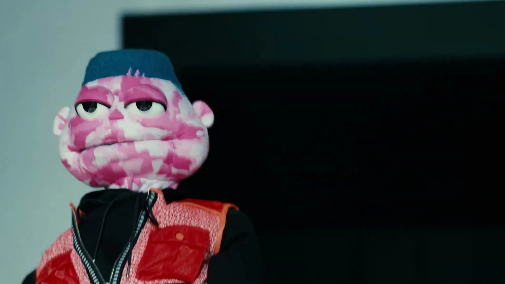 A puppet with pink camouflage skin, a blue-green mohawk, and a red vest looks around in an interior setting.