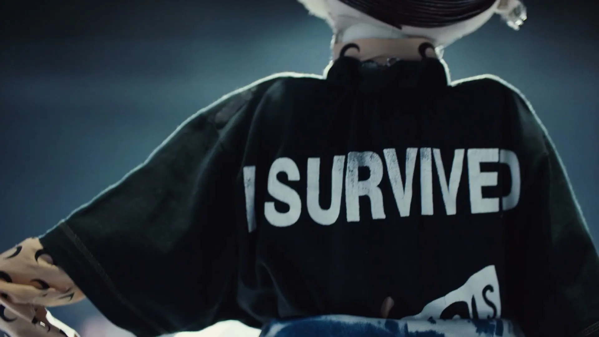 A person with an elaborate hairstyle wears a dark green jacket with "I SURVIVED" printed on the back, standing in a dark, indeterminate space.