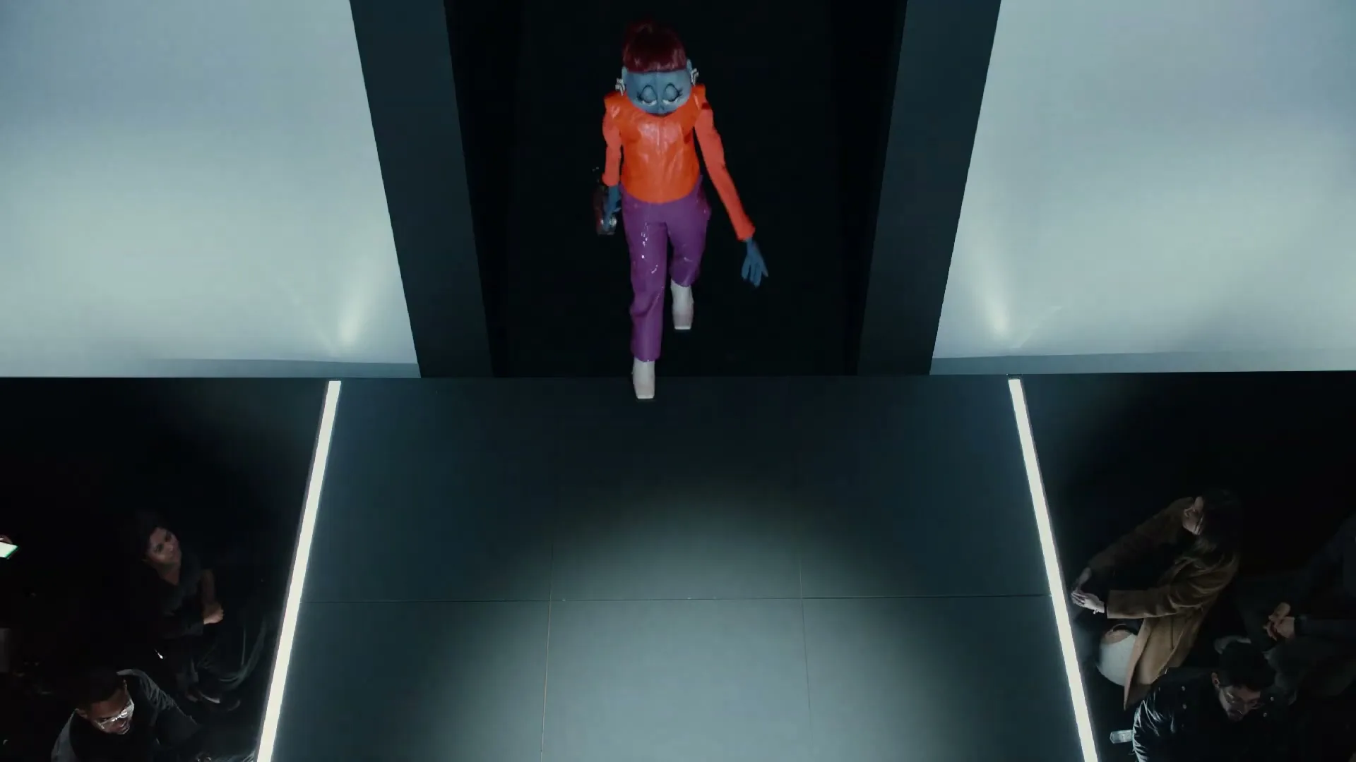 A blue-skinned, red-haired puppet in an orange jacket and purple pants walks down a runway, observed by a crowd of people on either side.