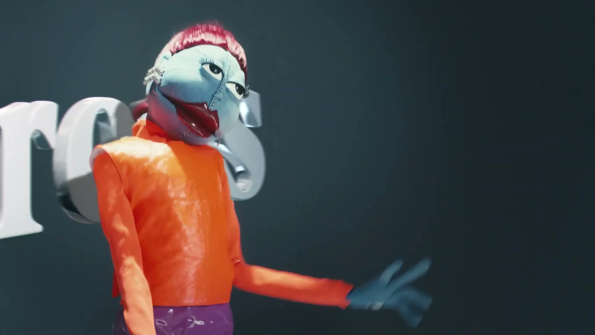 A blue-faced puppet with red hair and an orange top walks past large 3D letters spelling "Drops" against a dark background.