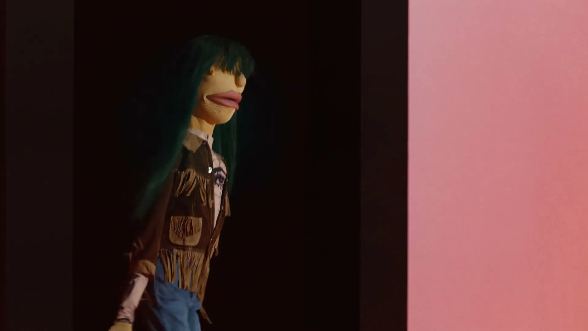A puppet with green hair, a brown fringed jacket, and blue jeans walks from left to right in a dark space with a pink wall.