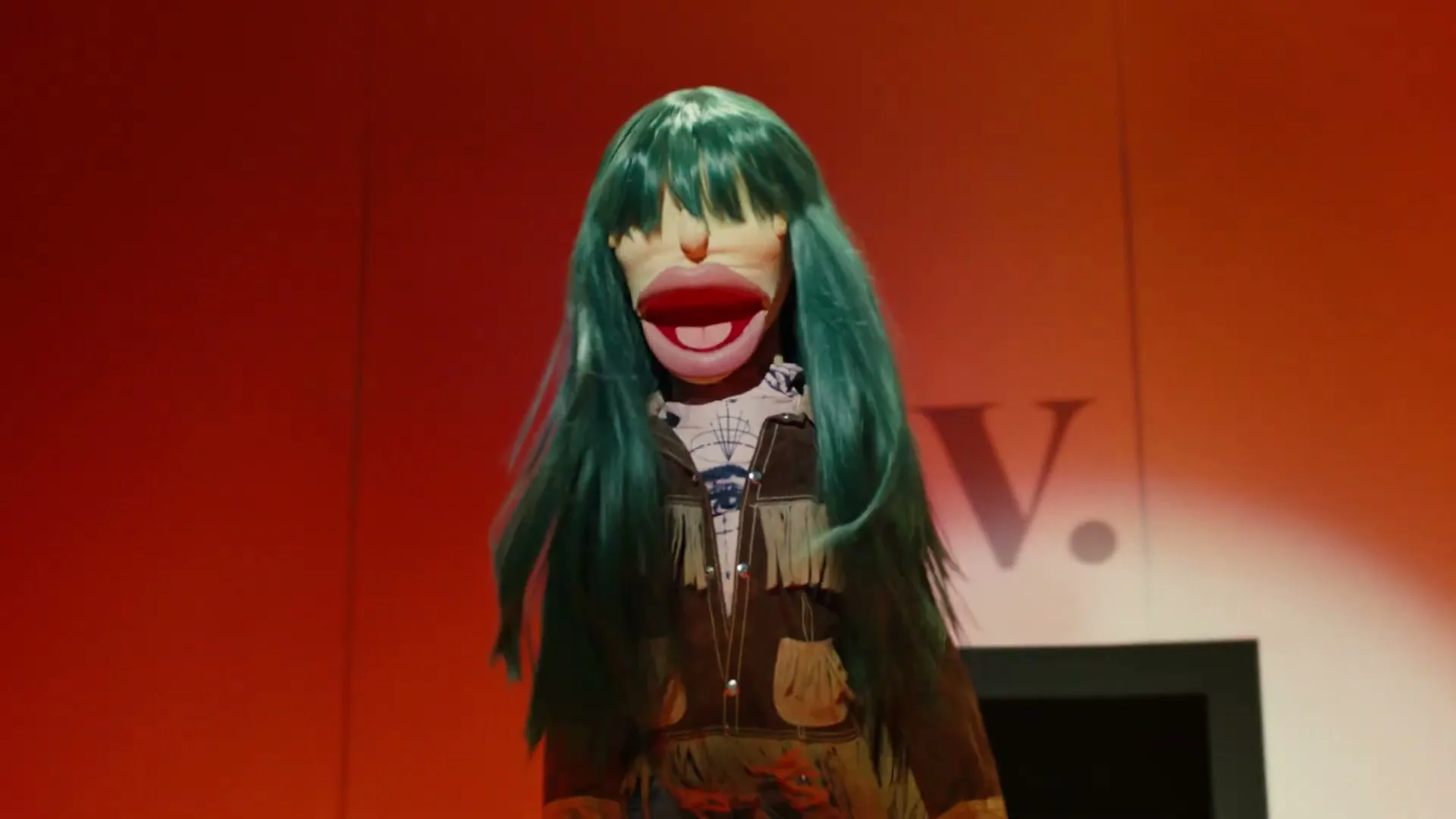 A puppet with long green hair and a fringed jacket stands in front of a wall with a large "V." logo, its mouth open.