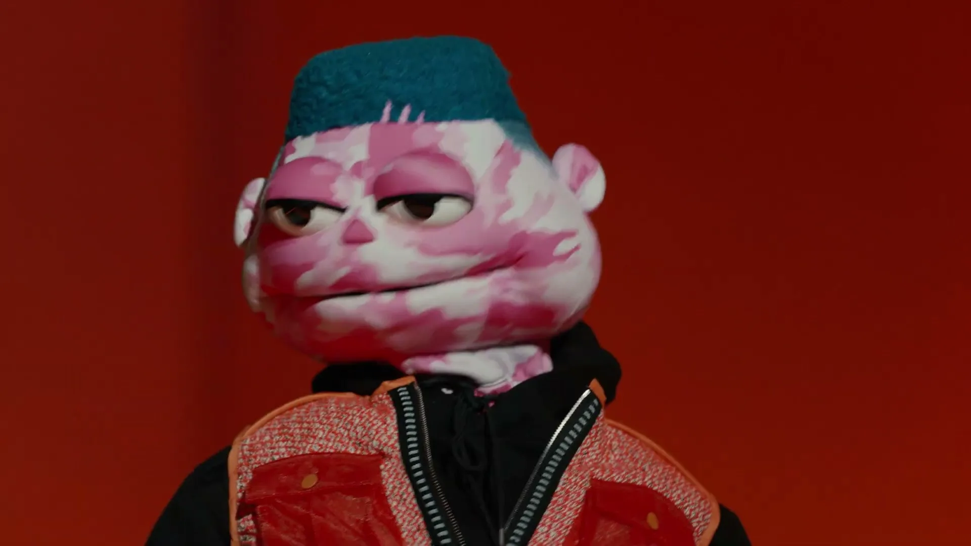 A puppet with pink camouflage skin, a blue mohawk, and a red vest looks around and smiles against a red background.