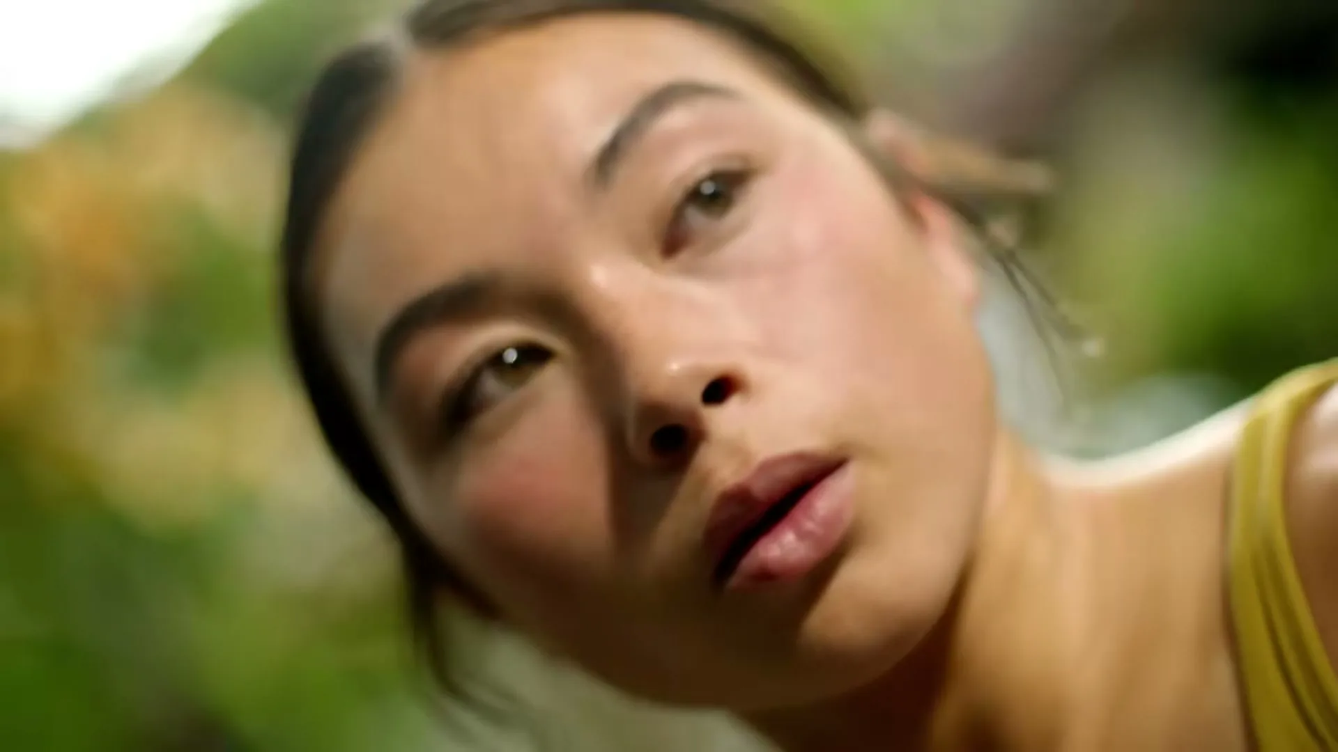 A young woman with brown hair and a yellow top looks up, then closes her eyes, in a soft focus outdoor setting.