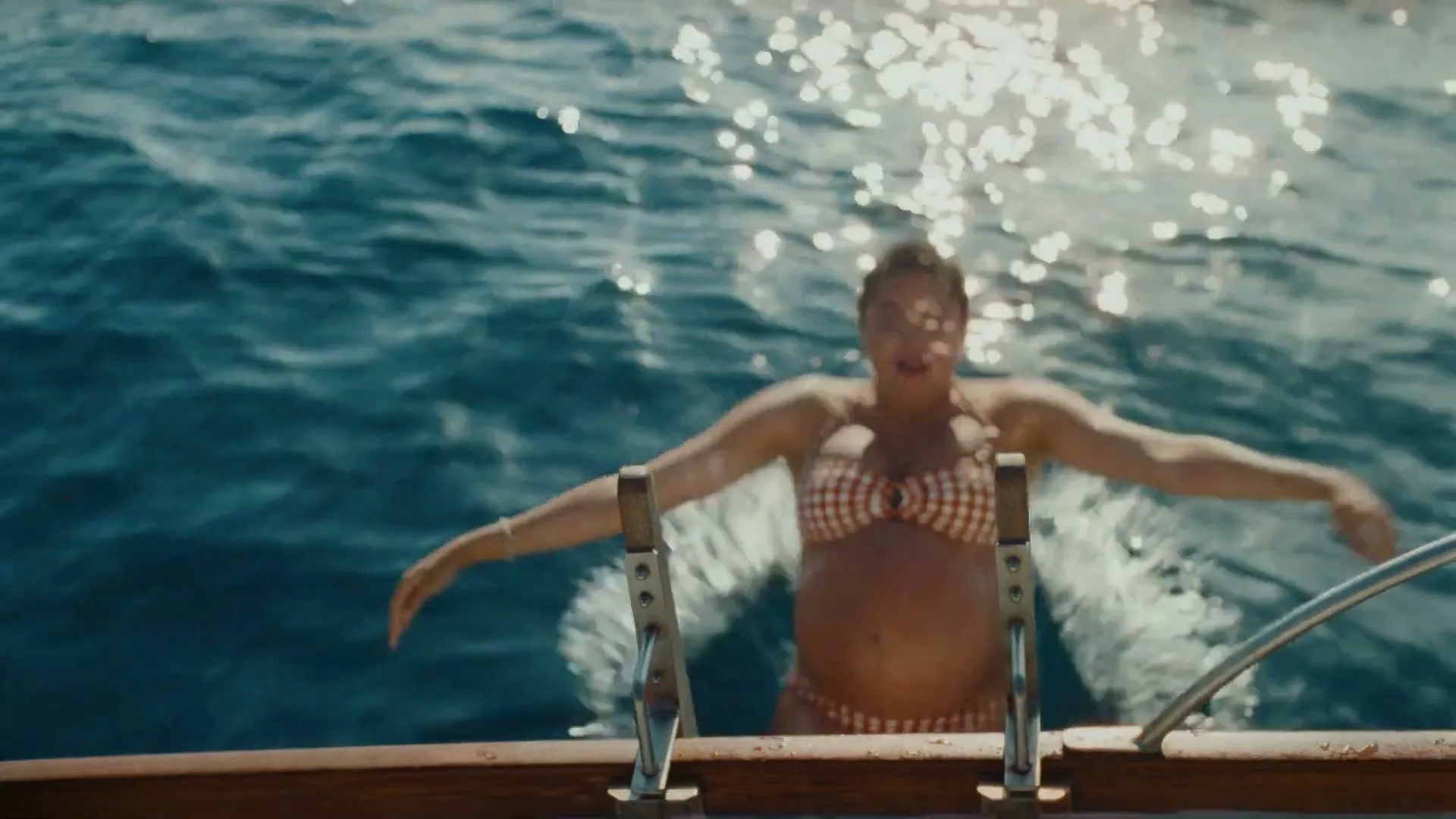A woman in a red and white gingham bikini jumps backward into the sparkling blue ocean from a boat.