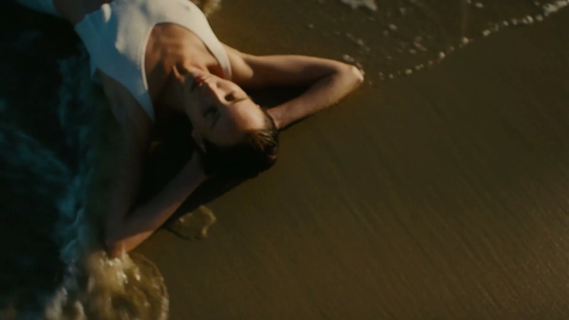 A woman in a white swimsuit lies on a sandy beach as gentle waves wash over her, captured from an overhead perspective.