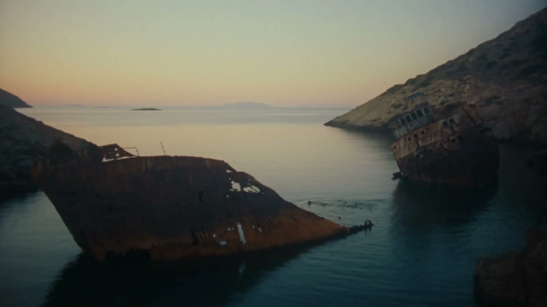 Two rusty shipwrecks are partially submerged in calm, blue water between rocky cliffs at dusk, with a small island visible in the distance.