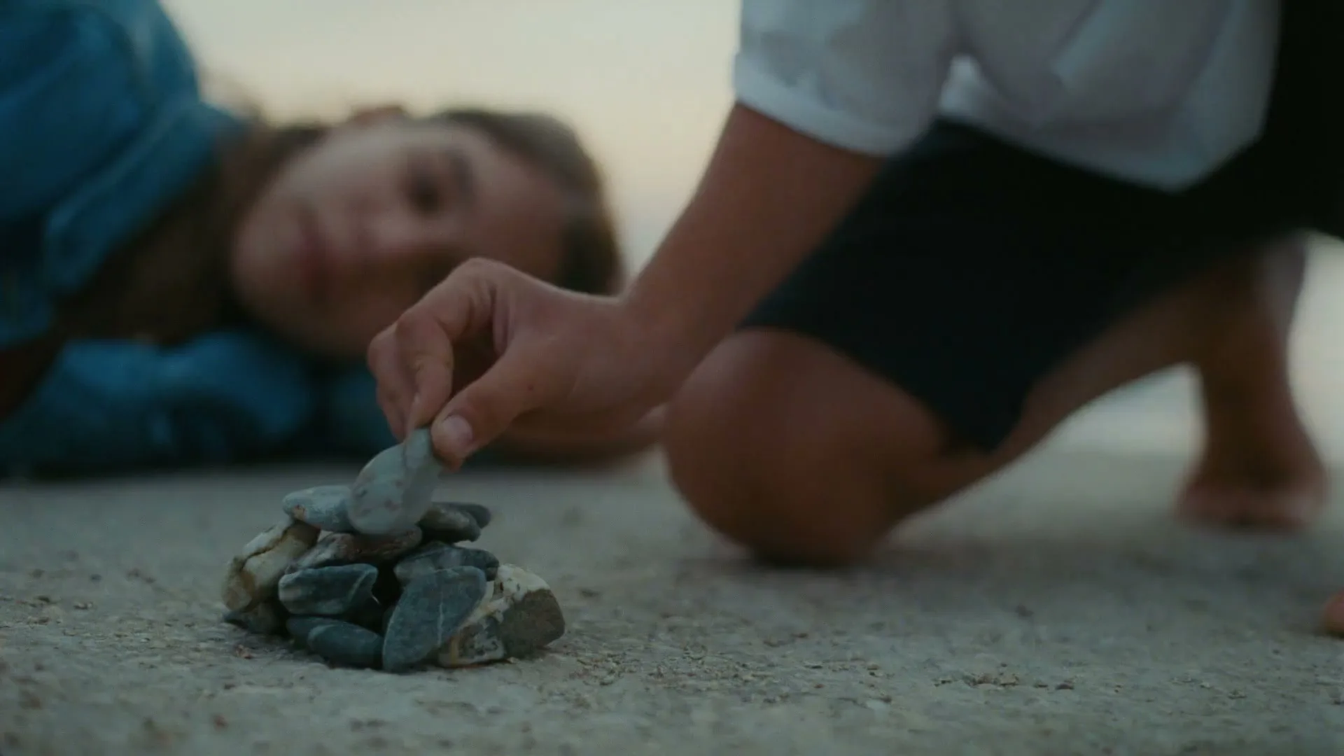 A young boy and girl are on a beach, the boy is building a small cairn of pebbles while the girl watches him, smiling.