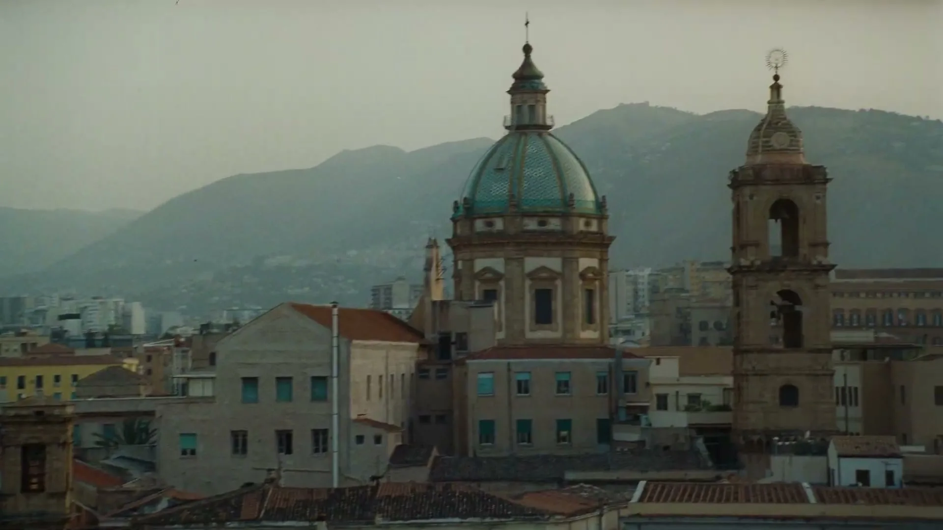 An establishing shot of a European city with a large dome and bell tower, with mountains in the background, under a hazy sky.