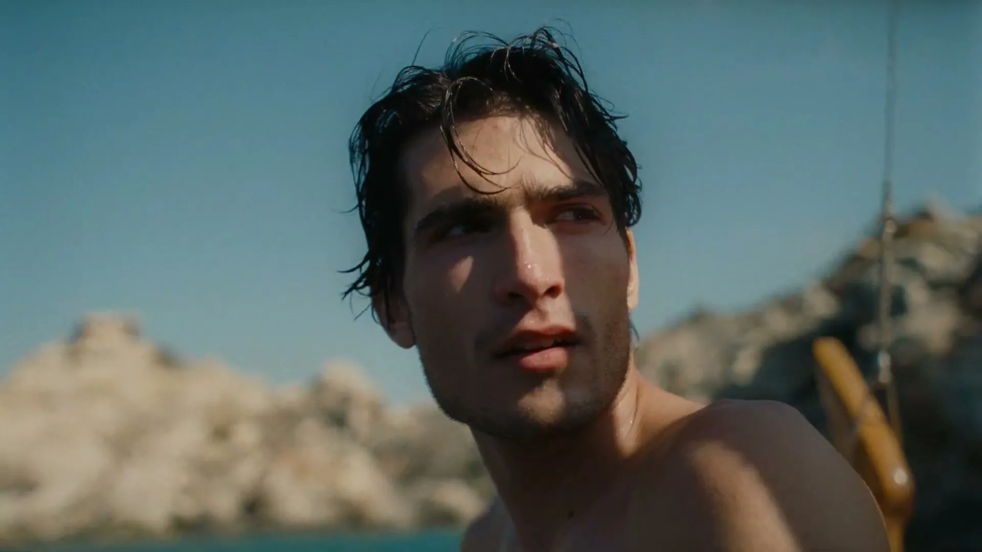 A young man with wet, dark hair and a slight stubble looks off-camera, with rocky cliffs and blue water in the background.