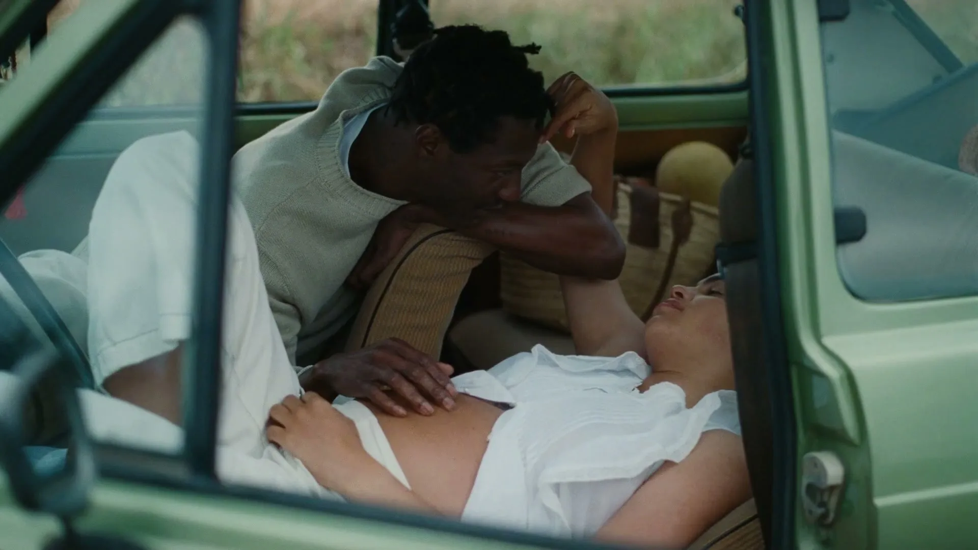 A man with dreadlocks leans over a woman lying down in the back of a light green car, touching her stomach.