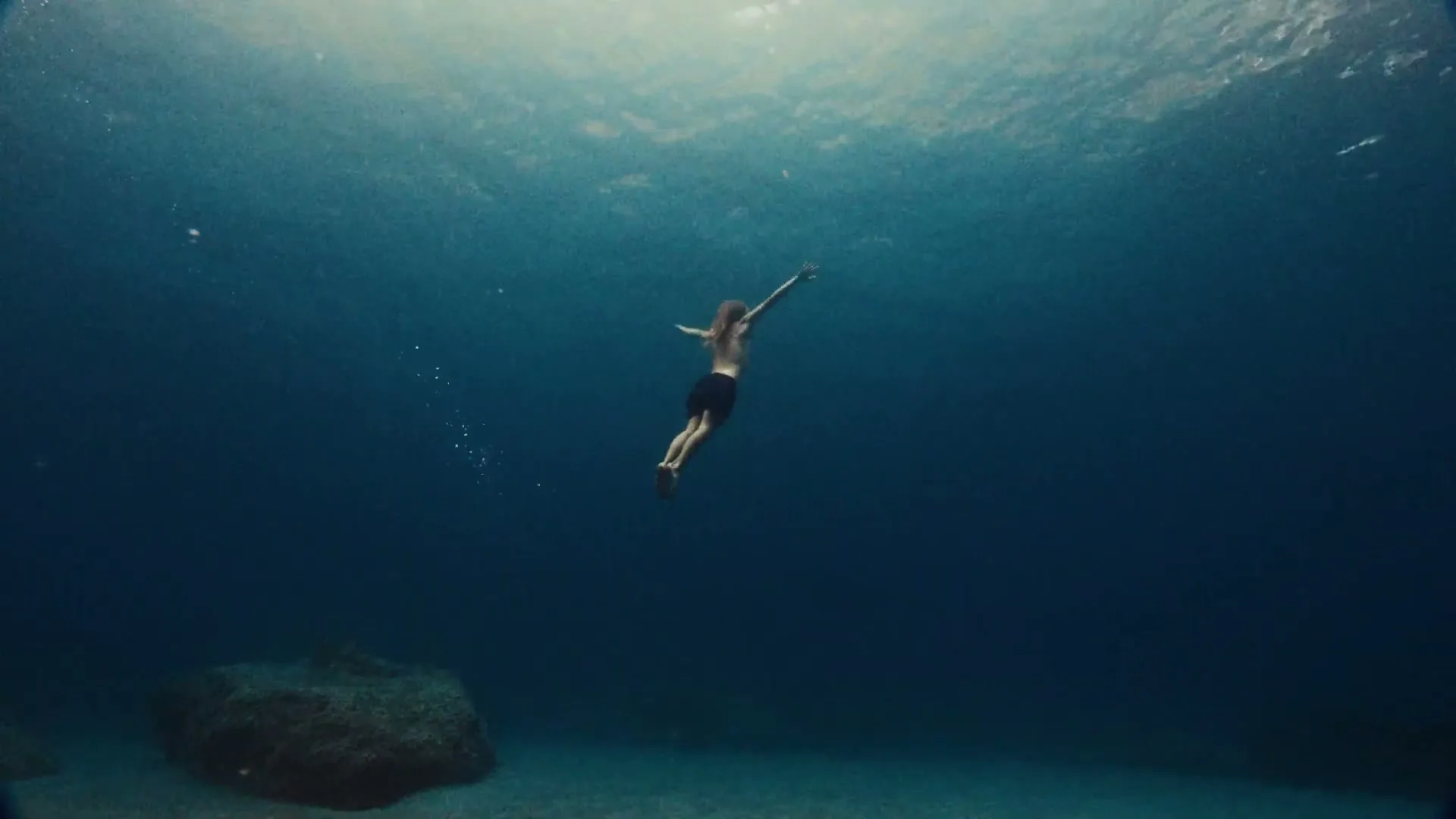 A person with long hair and dark shorts swims upwards in clear blue ocean water, with sunlight dappling from above.