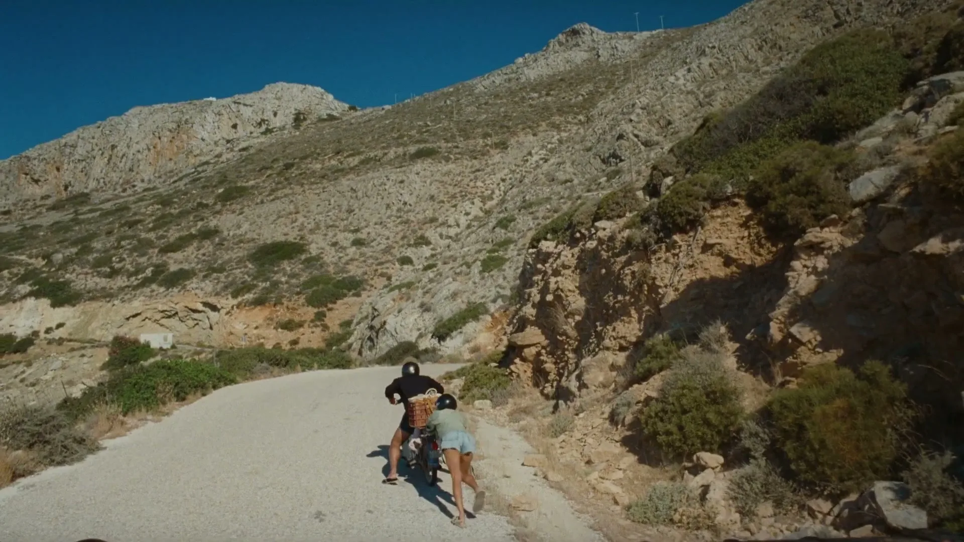 A man on a scooter and a woman pushing it up a winding road on a sunny day in a mountainous, arid landscape.