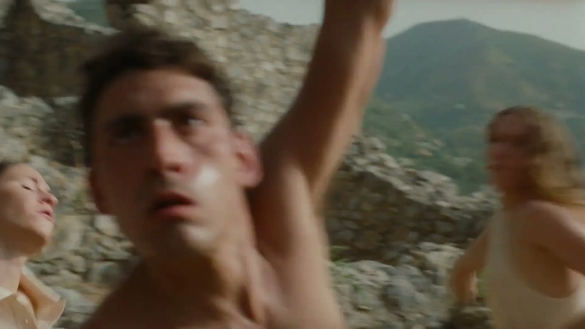 A group of young adults, some shirtless, are running and dancing energetically amidst ancient stone ruins on a sunny day, with mountains in the background.