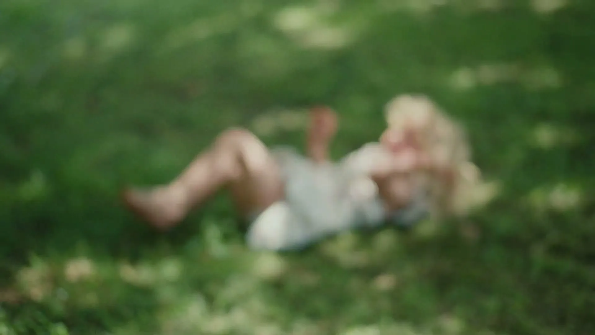 A blurry shot of a young girl with blonde hair, wearing a white dress, lying on green grass in a sun-dappled outdoor setting.