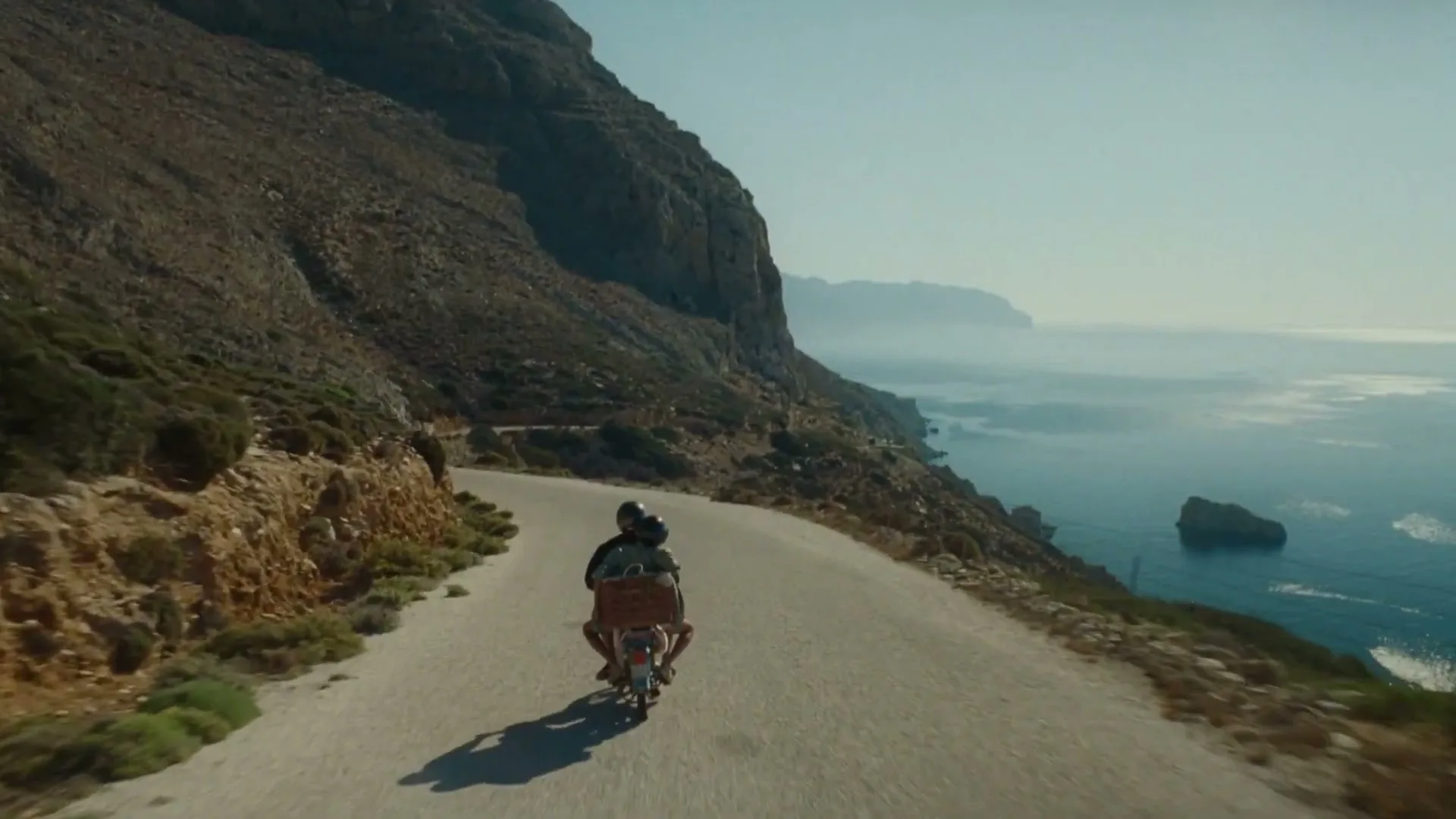 Two people on a scooter ride along a winding coastal road with a steep cliff on one side and the ocean on the other.