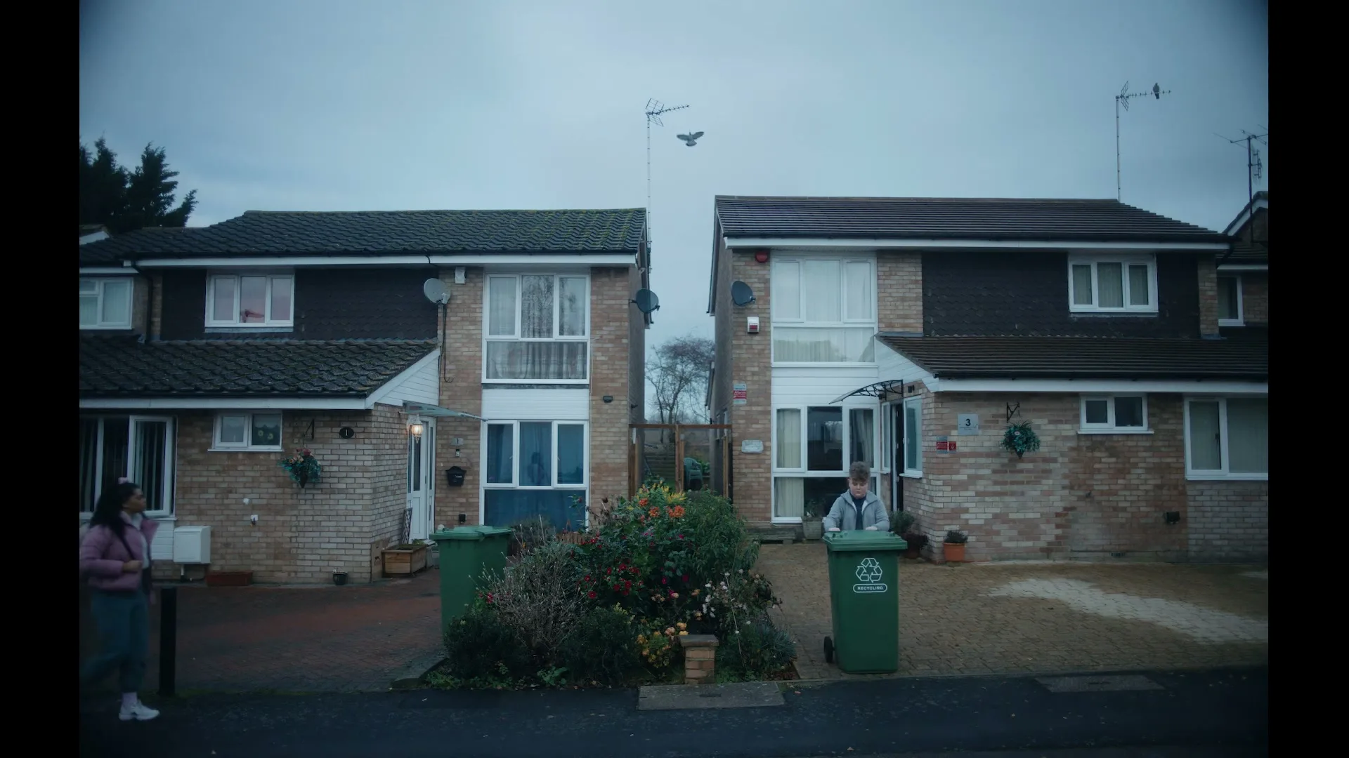 A young boy in a grey hoodie stands by a green recycling bin in front of a suburban house, as a girl walks past him on the sidewalk.