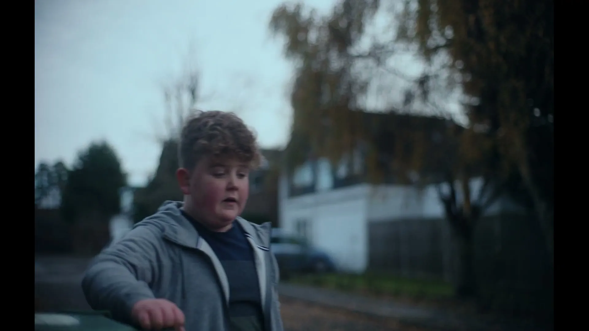 A young boy with curly hair leans on a car, looking off-camera with a neutral expression on a cloudy day in a suburban street.