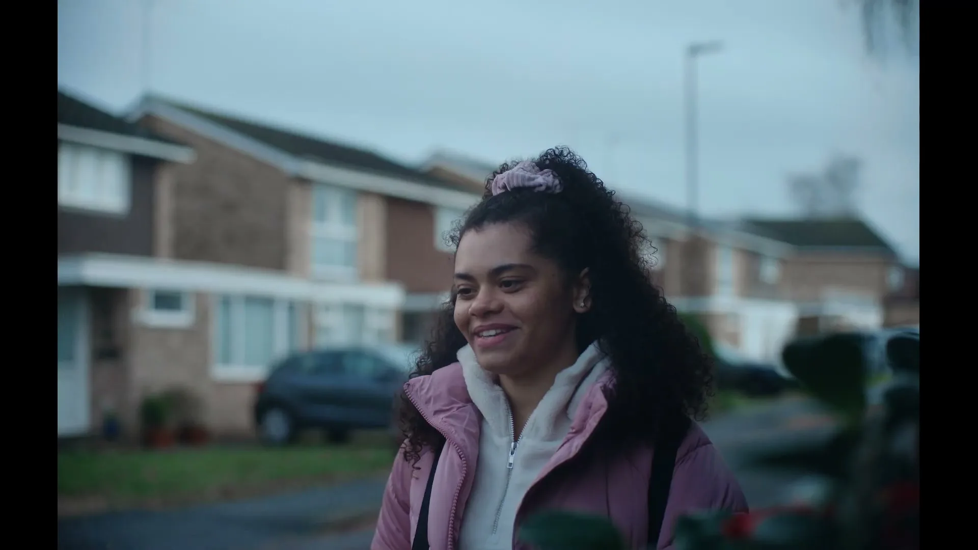 A young woman with curly hair and a pink jacket smiles while walking outdoors in a suburban neighborhood on a cloudy day.
