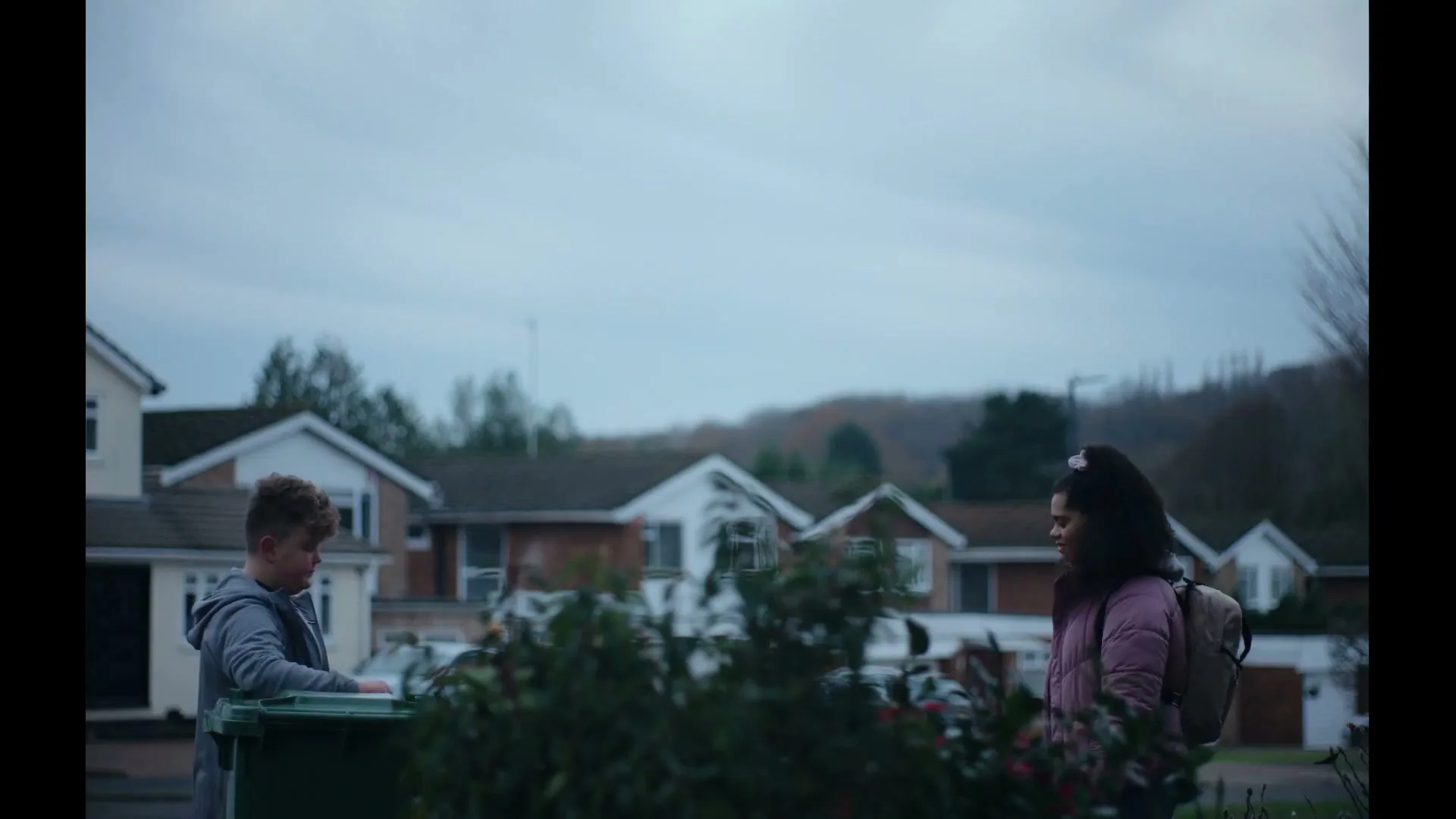 A young boy in a grey hoodie stands by a green recycling bin, looking at a woman with curly hair in a pink jacket and backpack, in a suburban street.