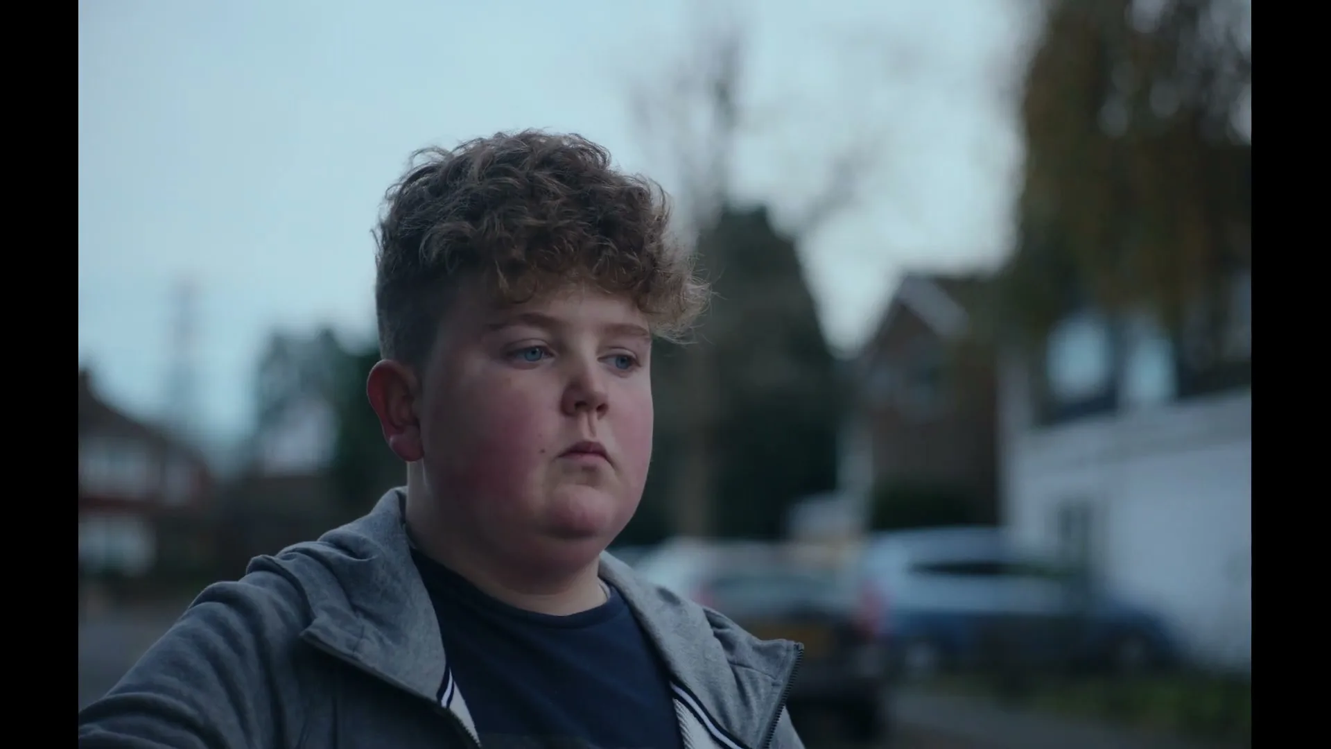 A young boy with curly hair and a grey hoodie stands outside, looking around with a slightly confused expression.