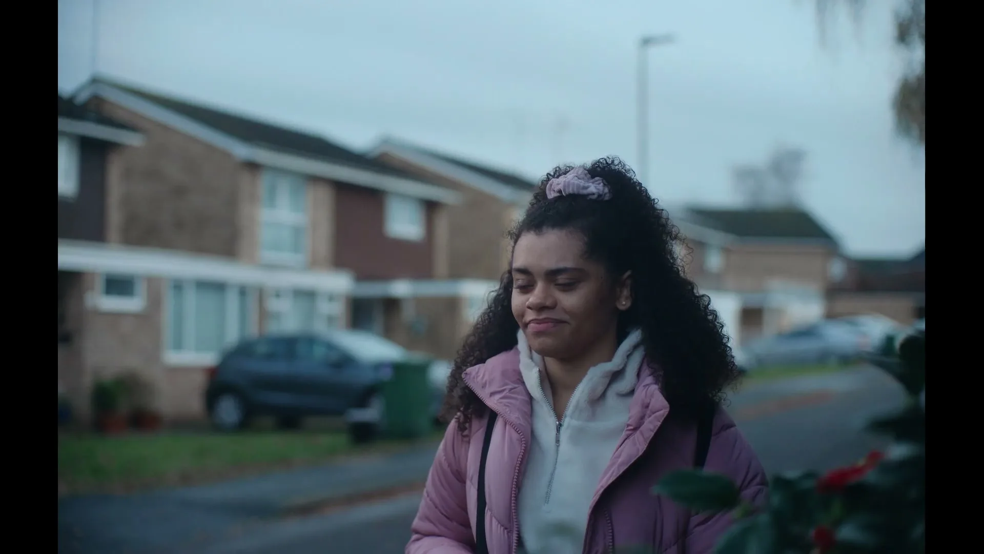A young woman with curly hair, wearing a pink puffer jacket and a backpack, walks down a suburban street looking thoughtful.