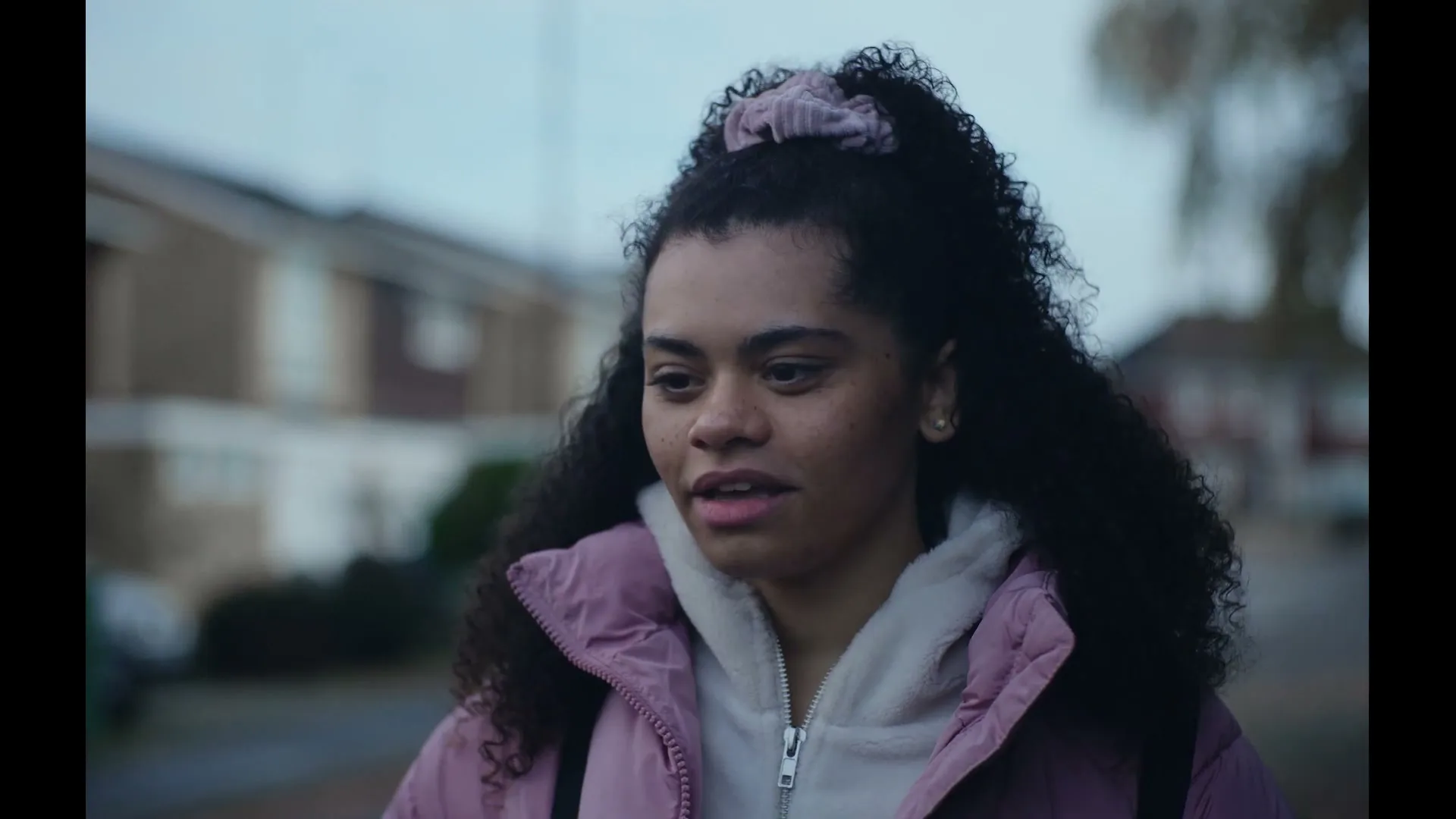 A young woman with curly hair and a pink jacket looks around with a slightly concerned expression on a suburban street.
