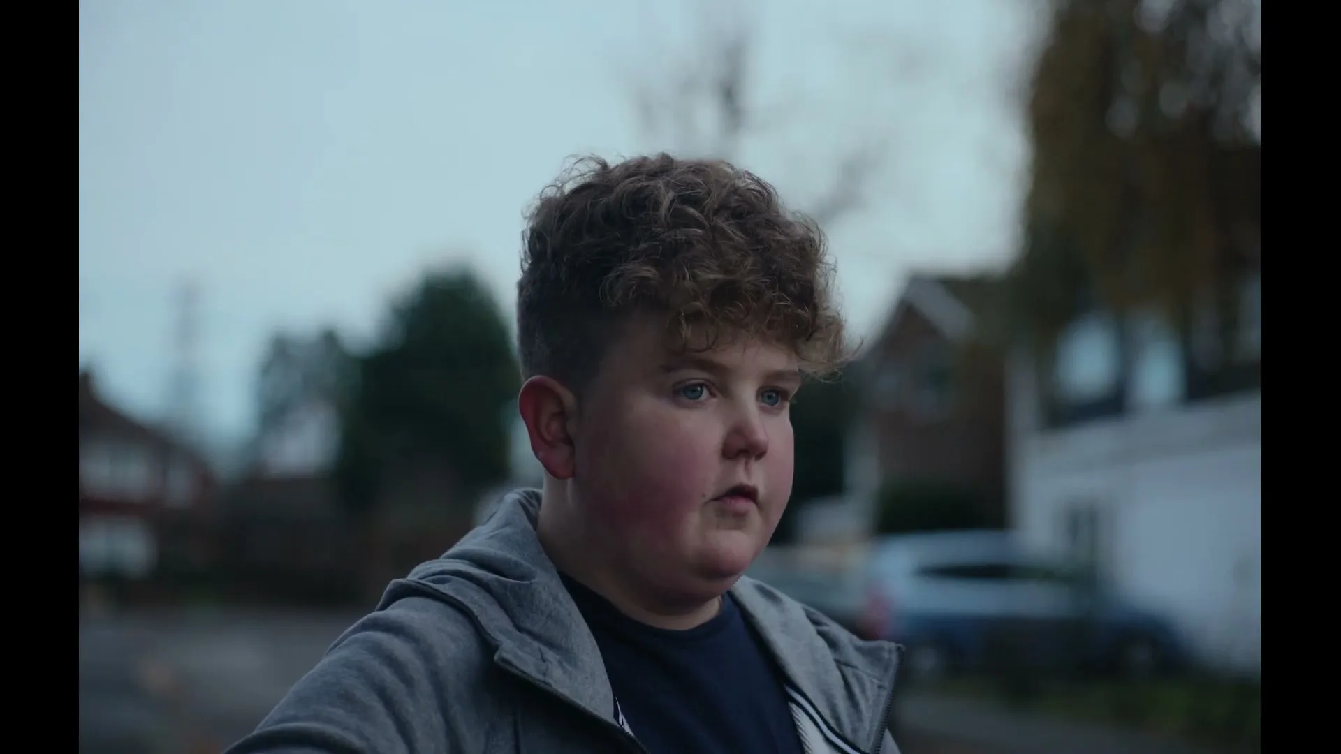 A young boy with curly hair and a grey hoodie looks down, then up, then down again, with a suburban street in the background on a cloudy day.