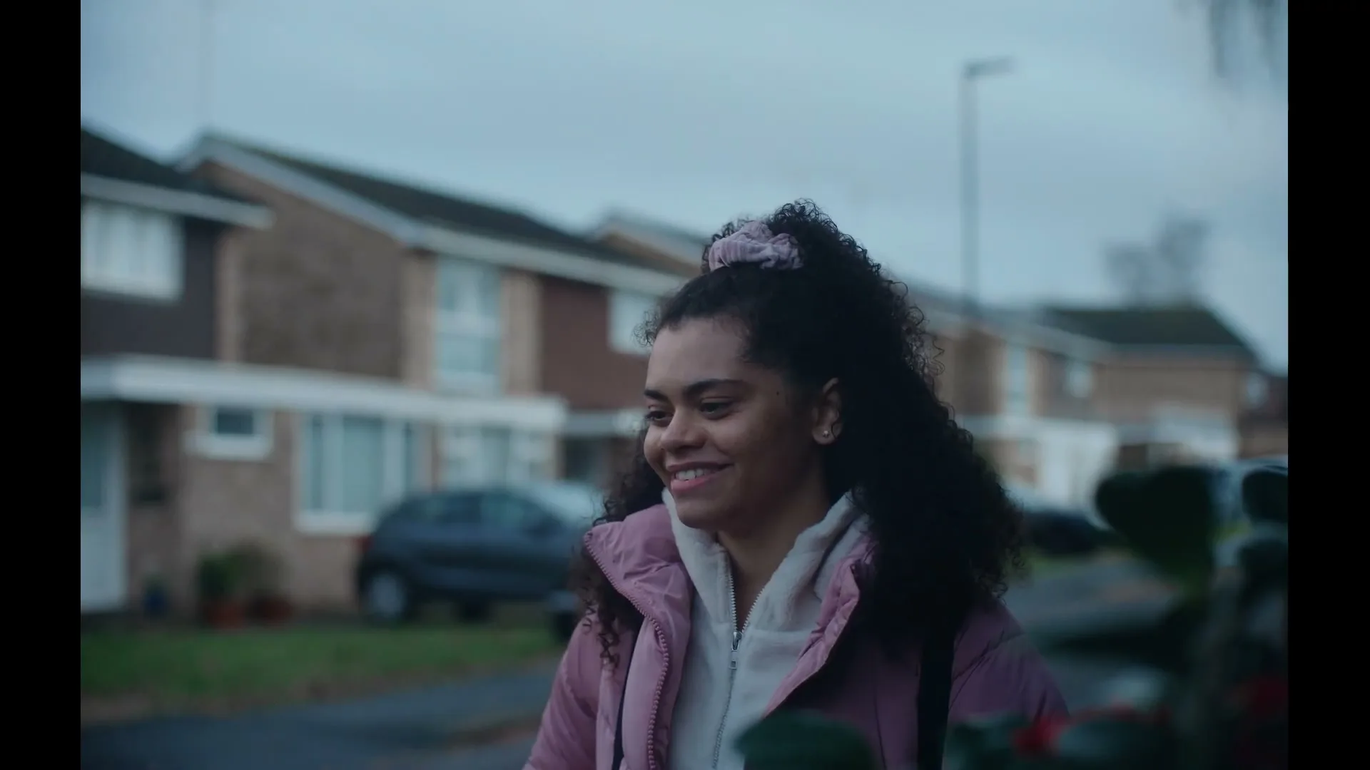 A young woman with curly hair in a pink jacket walks down a suburban street, smiling as she looks off-camera.