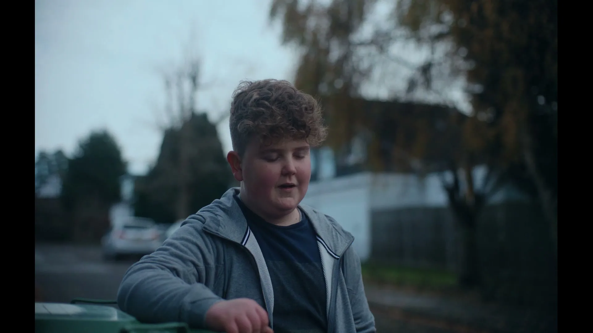 A young boy with curly hair leans on a green bin, looking downcast on a cloudy day in a suburban street.