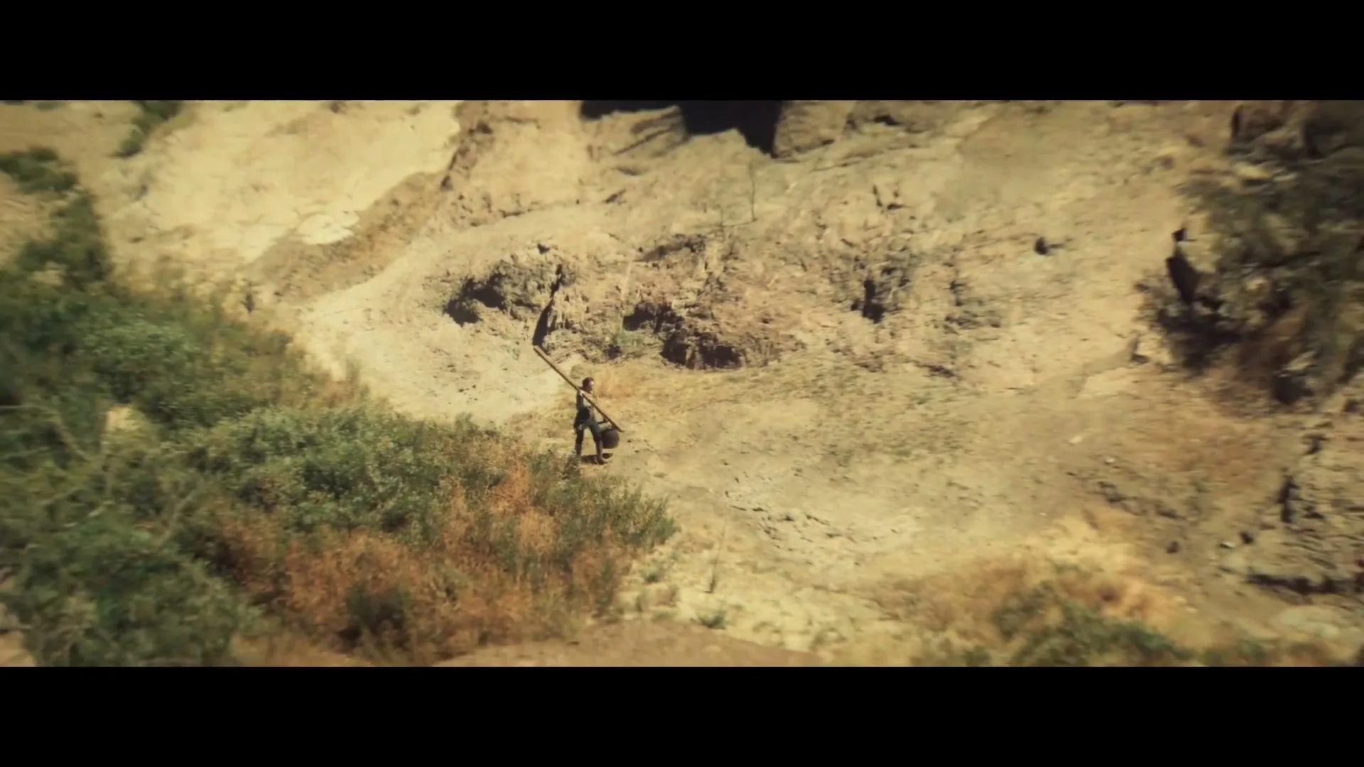 A person carries a long wooden beam and a bucket across a dry, rocky landscape, walking away from the camera as the shot pulls back to reveal a vast canyon.