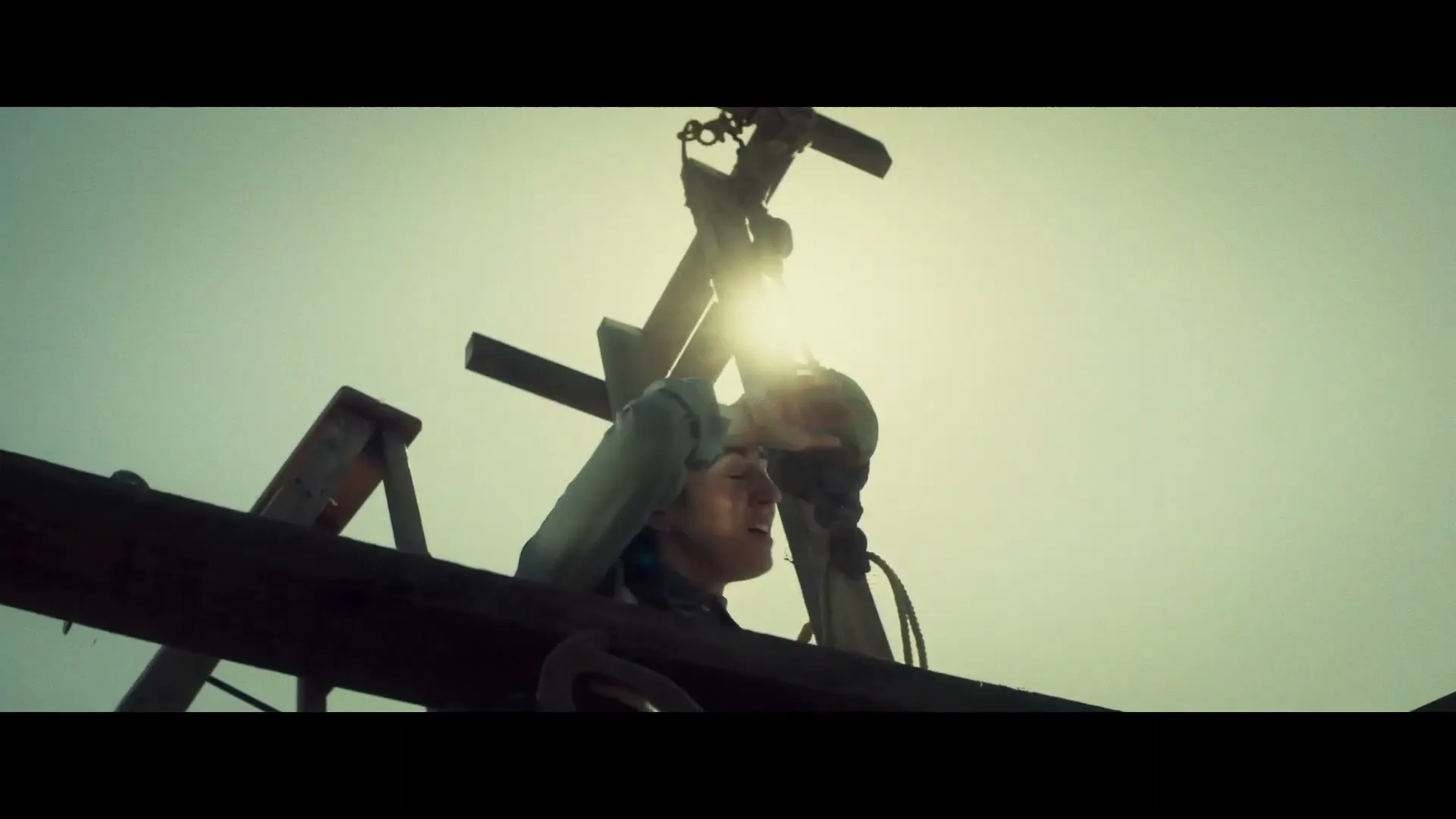 A person in work clothes looks up, shielding their eyes from the bright sun, while working on a wooden structure outdoors.