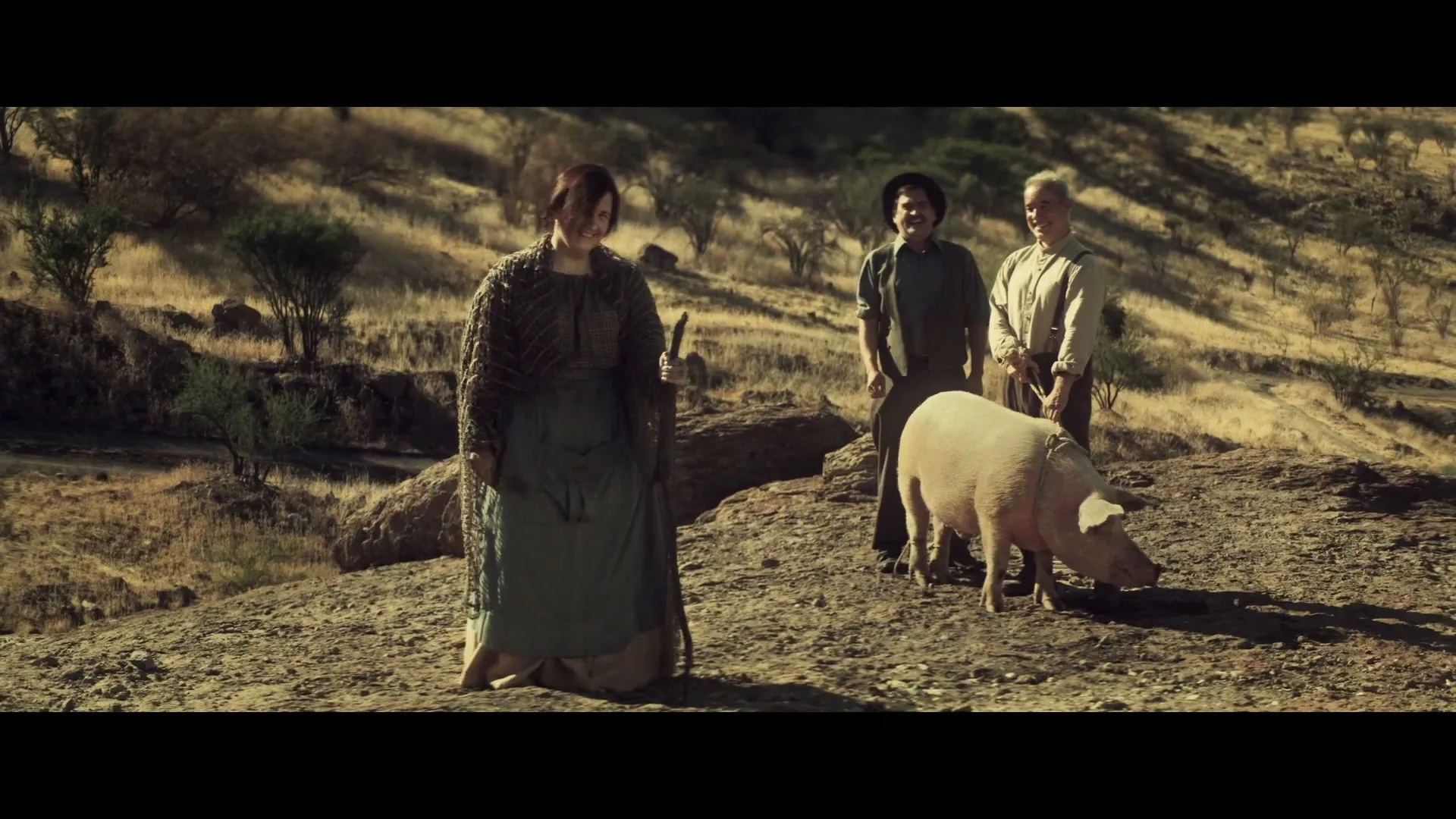 A woman in old-fashioned clothing stands with a stick, looking at two men and a pig on a dry, hilly landscape under a bright sky.