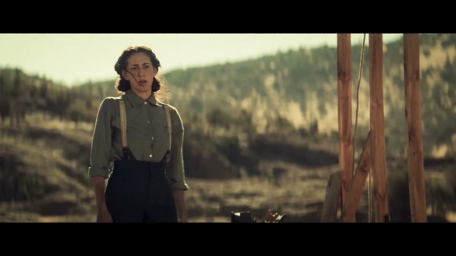 A woman with dark hair in a button-up shirt and suspenders stands in a dry, mountainous landscape, looking concerned.