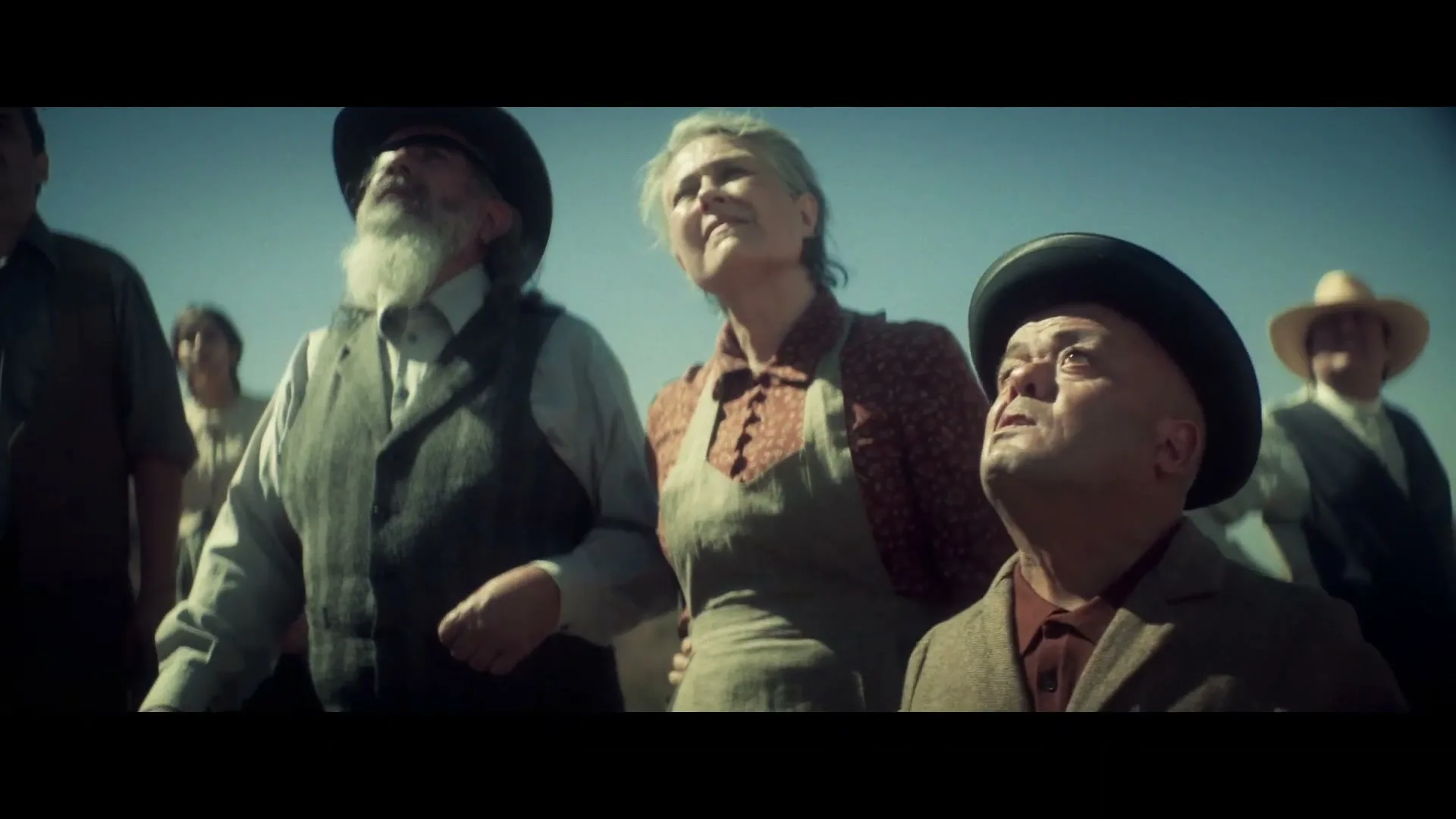 A group of early 20th-century settlers, including a bearded man, a woman in an apron, and a shorter man in a bowler hat, look up at the sky in an open field.