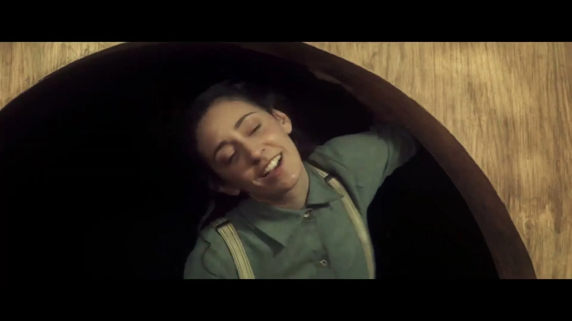 A young woman with dark hair and a green shirt with suspenders smiles and laughs while looking out from a dark, circular opening in a wooden structure.