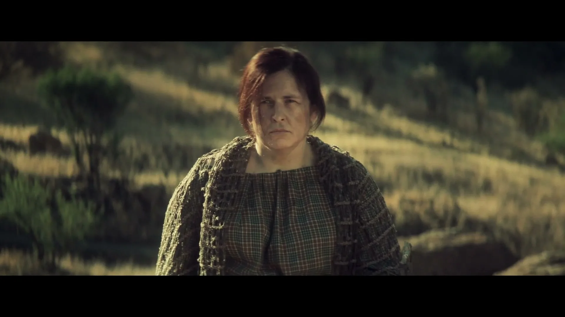 A determined woman with dark hair and a plaid dress stands in a field, looking directly at the camera with a serious expression.